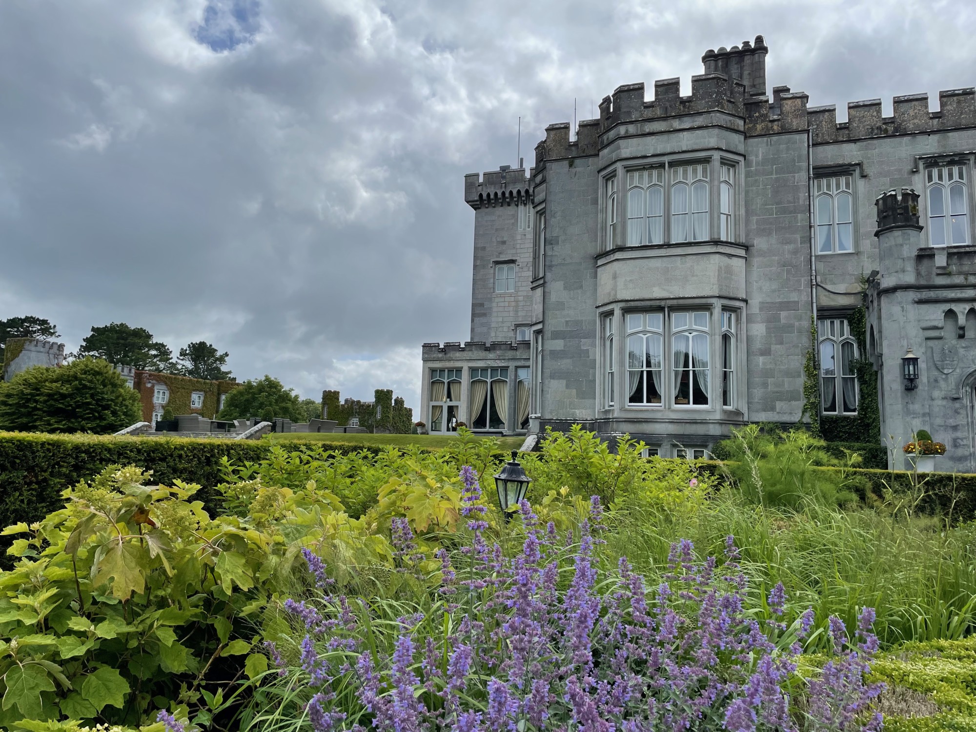 Grey castle surrounded by green garden with purple flowers.