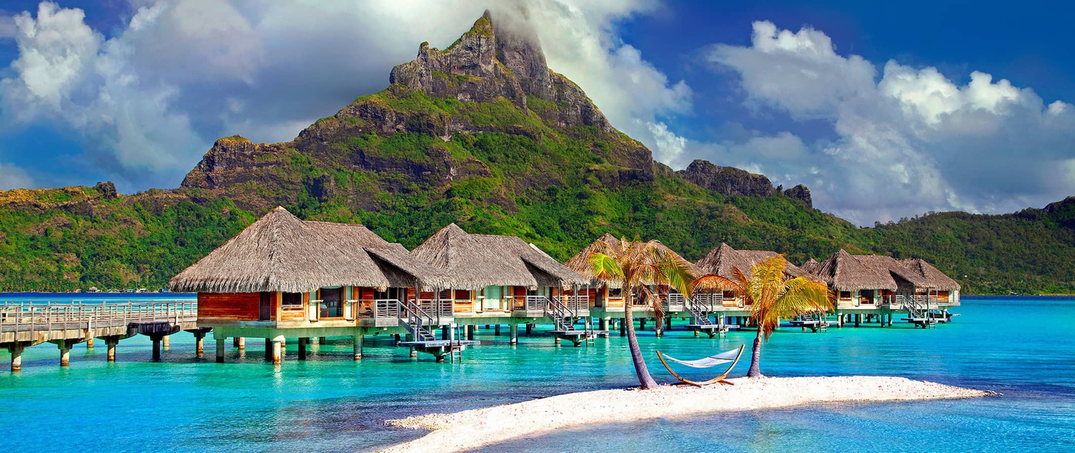 Bungalows over blue water during the daytime with a mountain peak in the distance