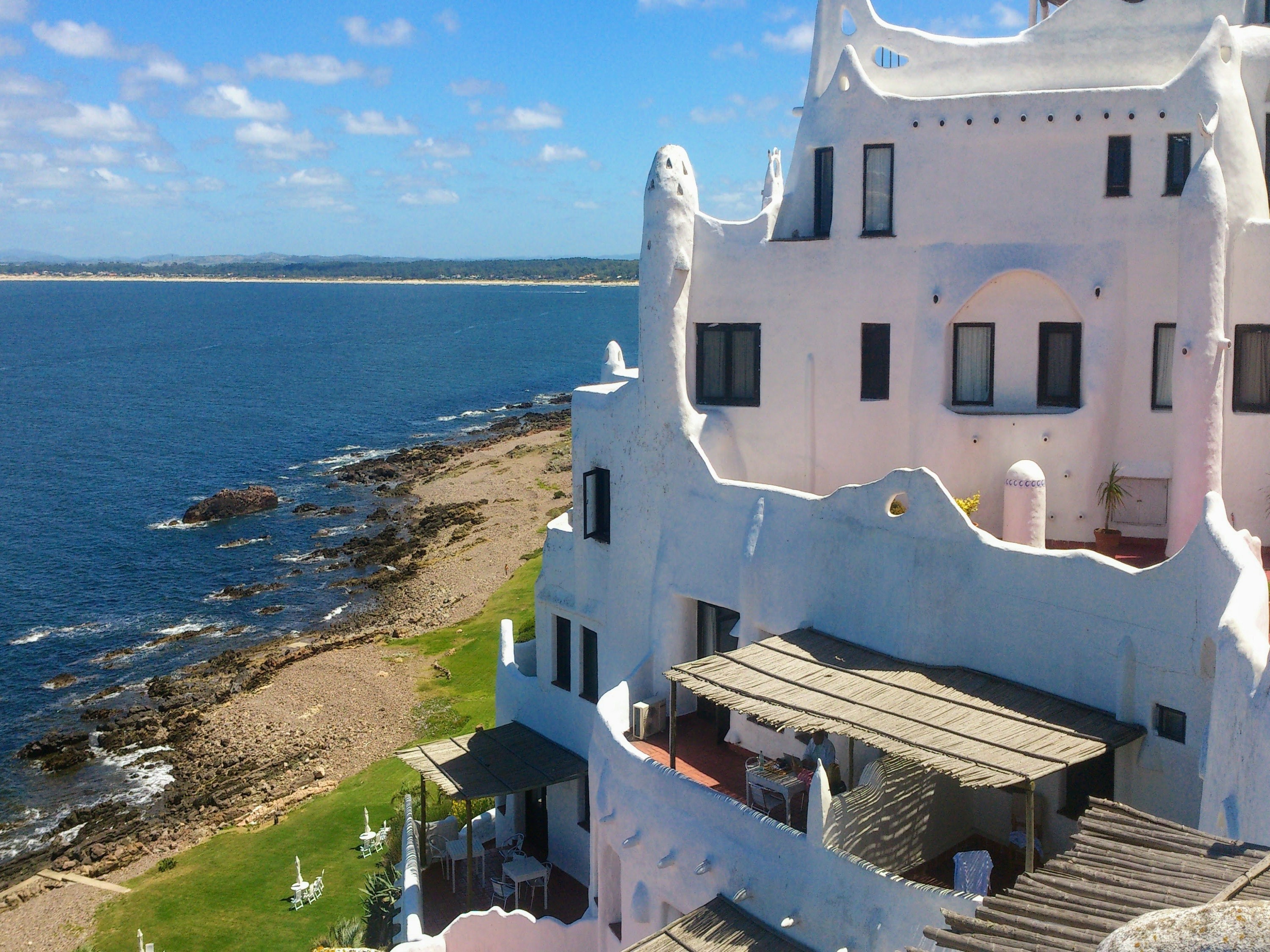 The image captures a unique white building with curved walls and irregular windows, set against a rocky shoreline and calm blue sea under a clear sky.