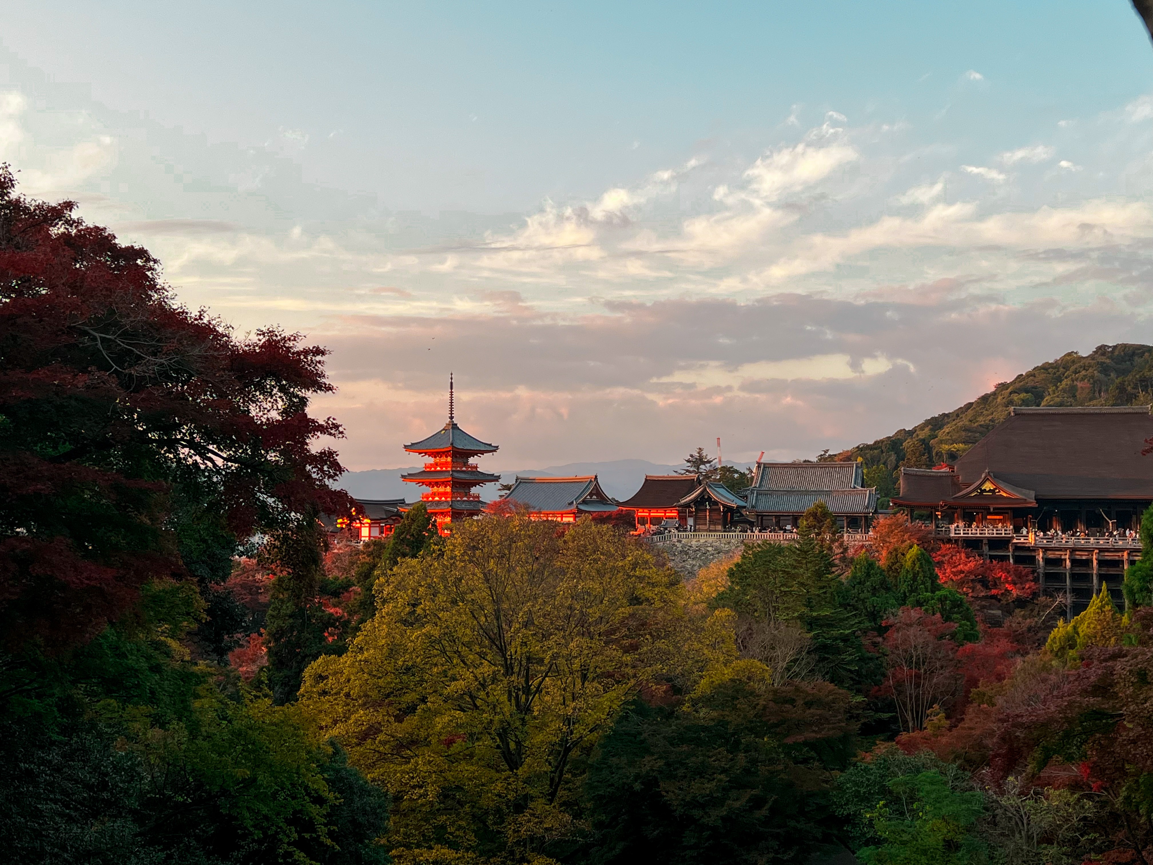 A picturesque Japanese temple complex with a red and white pagoda, surrounded by autumn-colored trees under a soft cloudy sky.