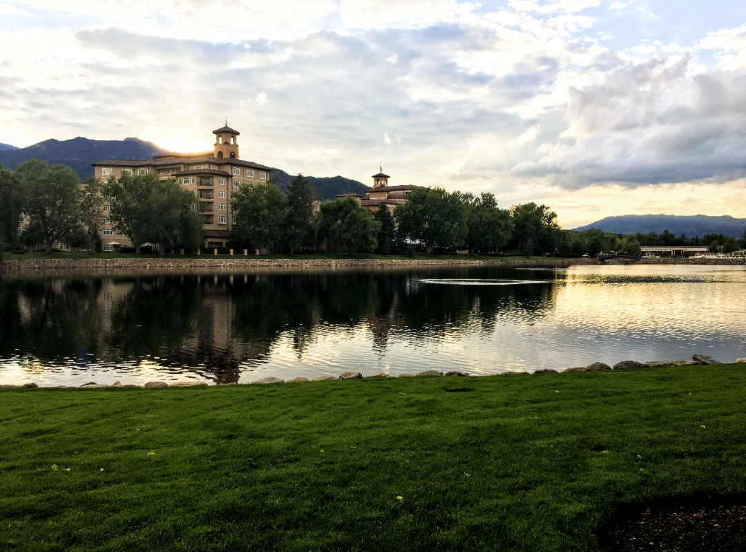 A view of green grass, a reflective pond and a beautiful building surrounded by trees and mountains in the background. There is a golden sunset reflecting over the water.