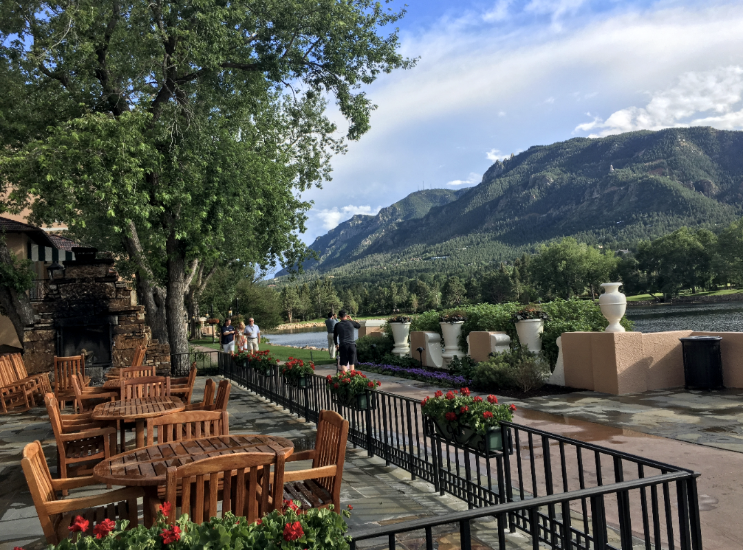 A picture of an outdoor patio with wooden tables and chairs, a sidewalk, planters, trees and a stunning view of a mountain range toward the right side of the image.