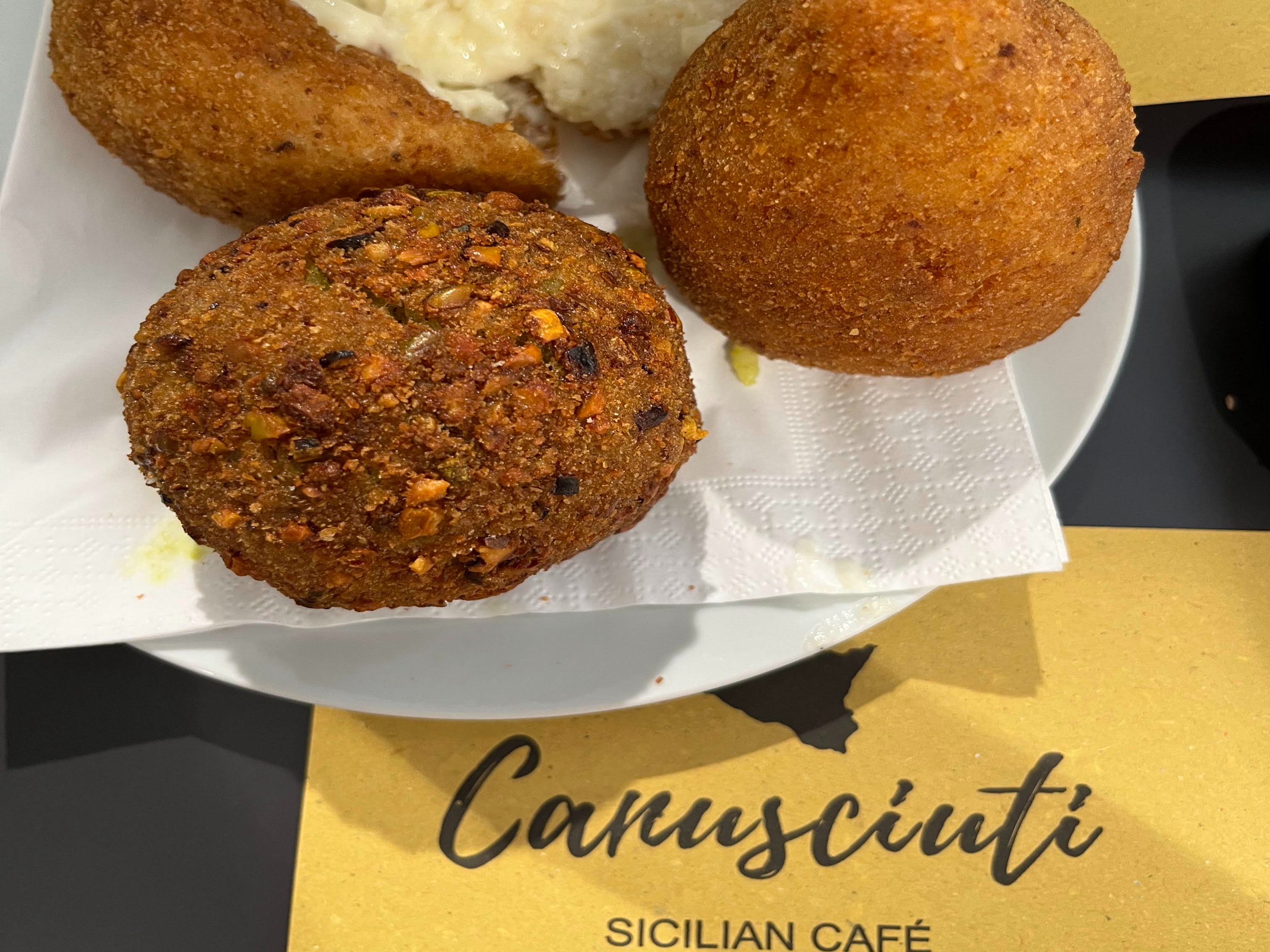 The image displays a variety of Sicilian fried snacks on a plate, with the name “Cannscutti Sicilian Café” visible, suggesting a culinary delight.