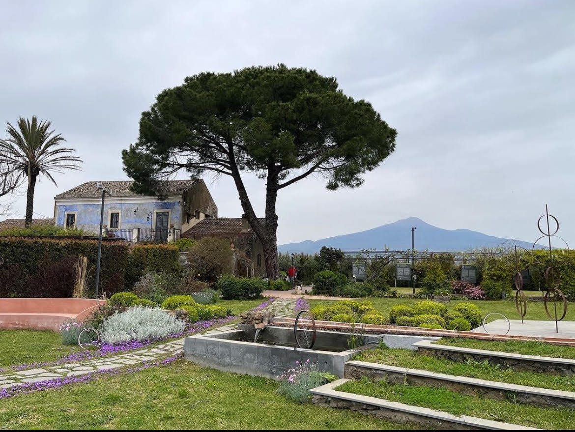The image offers a scenic outdoor view with a large tree, a house, Mount Vesuvius in the background, and sculptures in a garden.