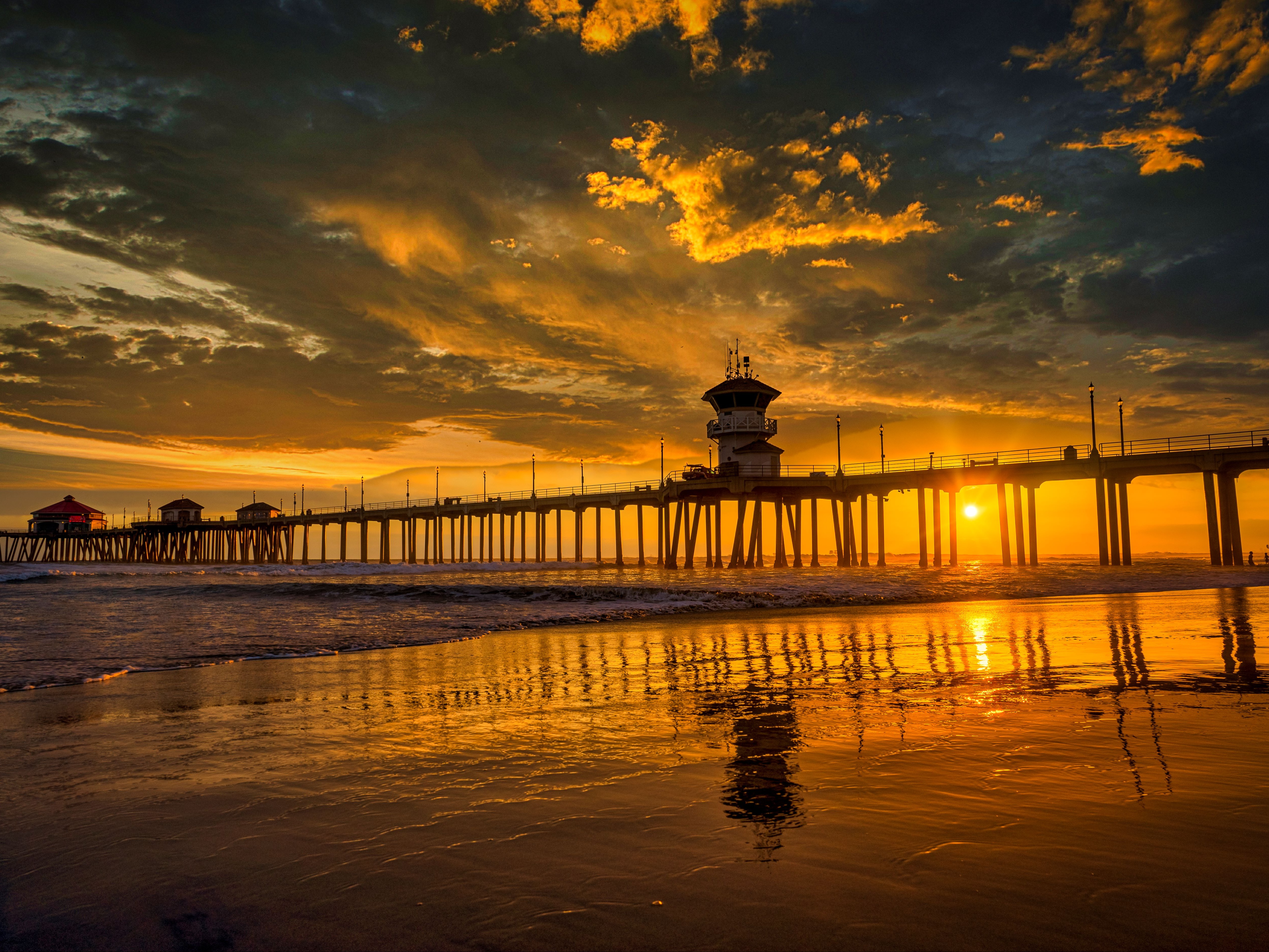 The image depicts a tranquil sunset at a beach, with a pier and lighthouse casting reflections on the wet sand.