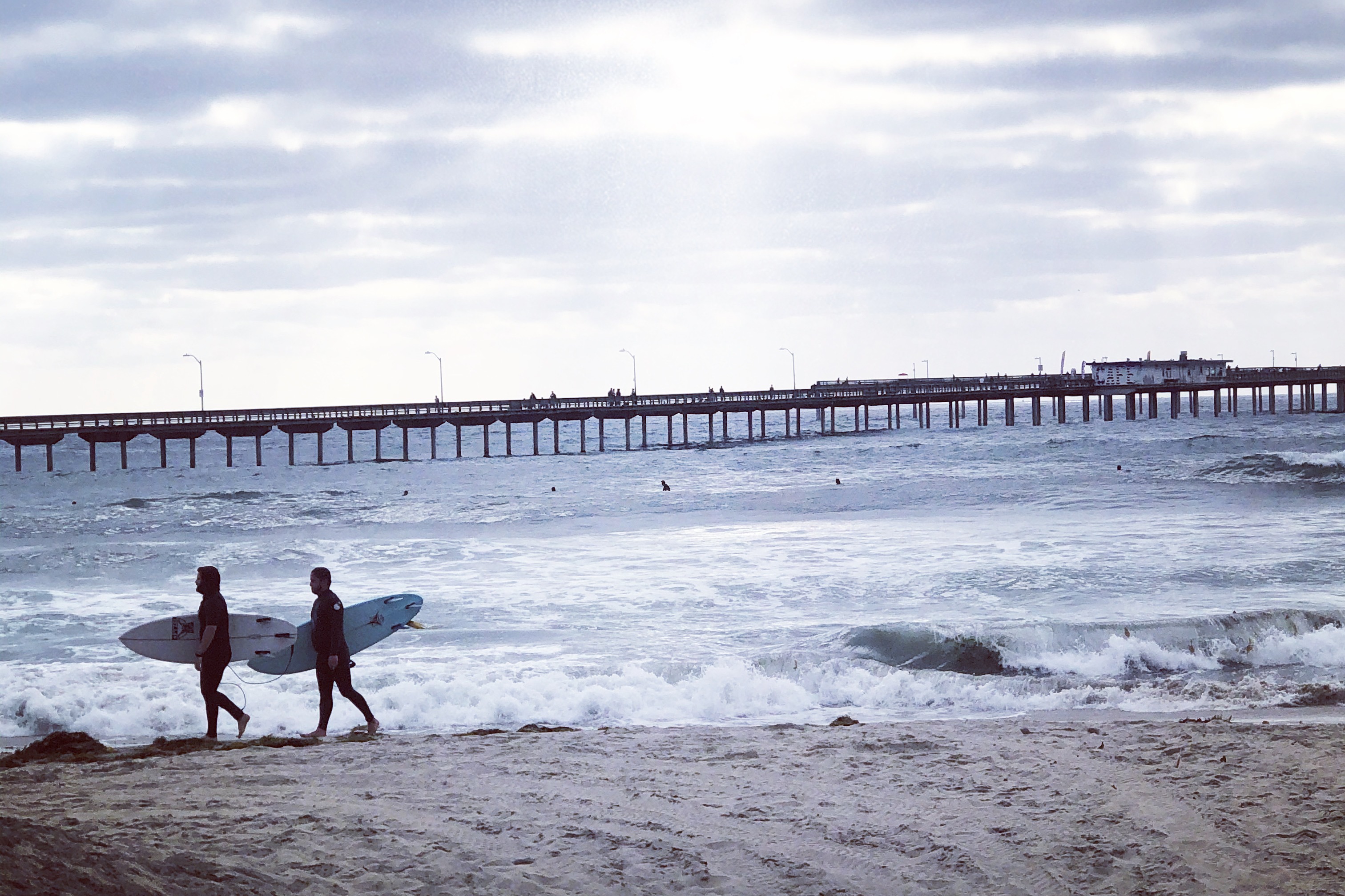 Two people walking on the beach carrying surf boards during a cloudy day