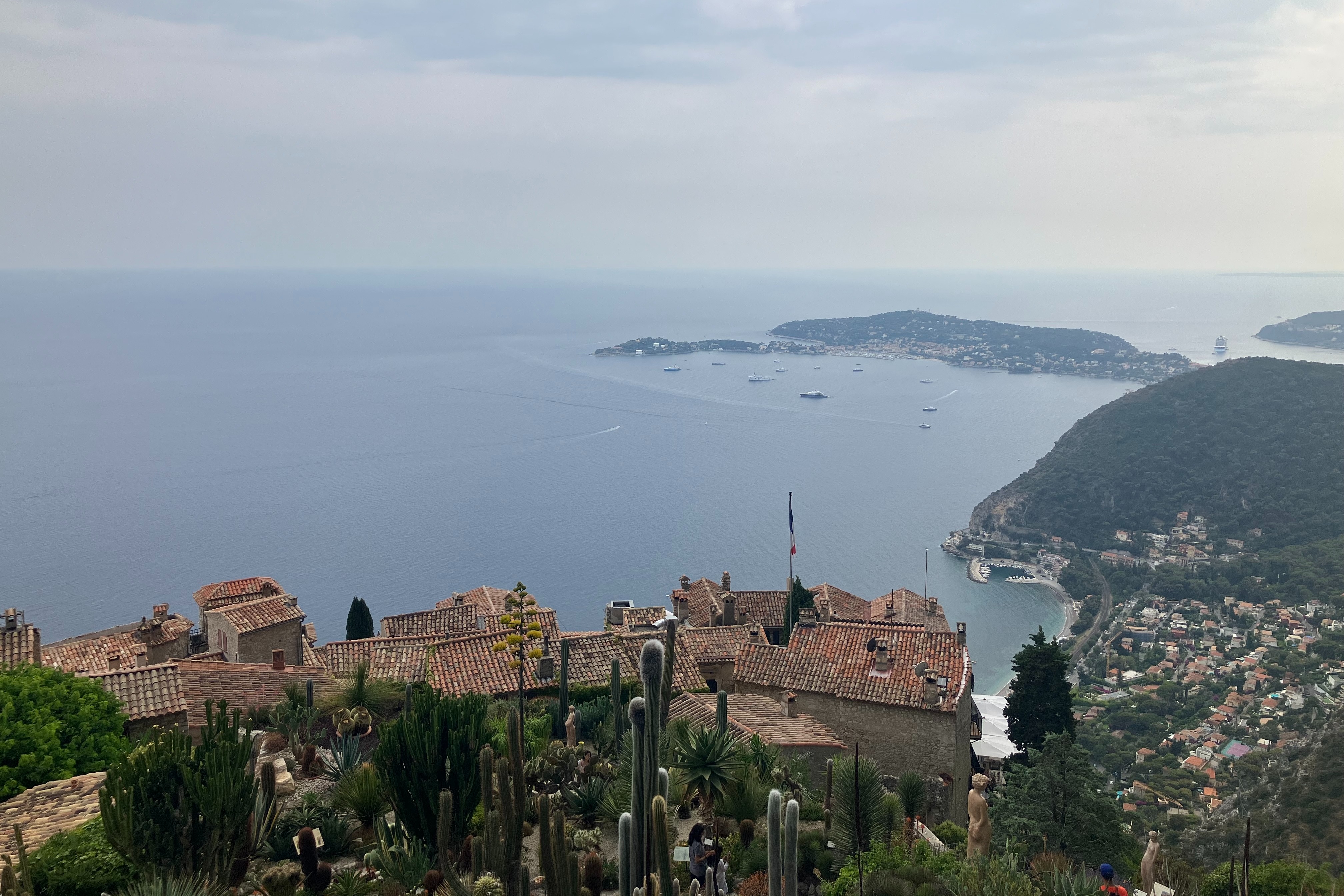 A picturesque coastal vista, showcasing terracotta rooftops amidst lush greenery, with a tranquil sea and boats under a soft, hazy sky.