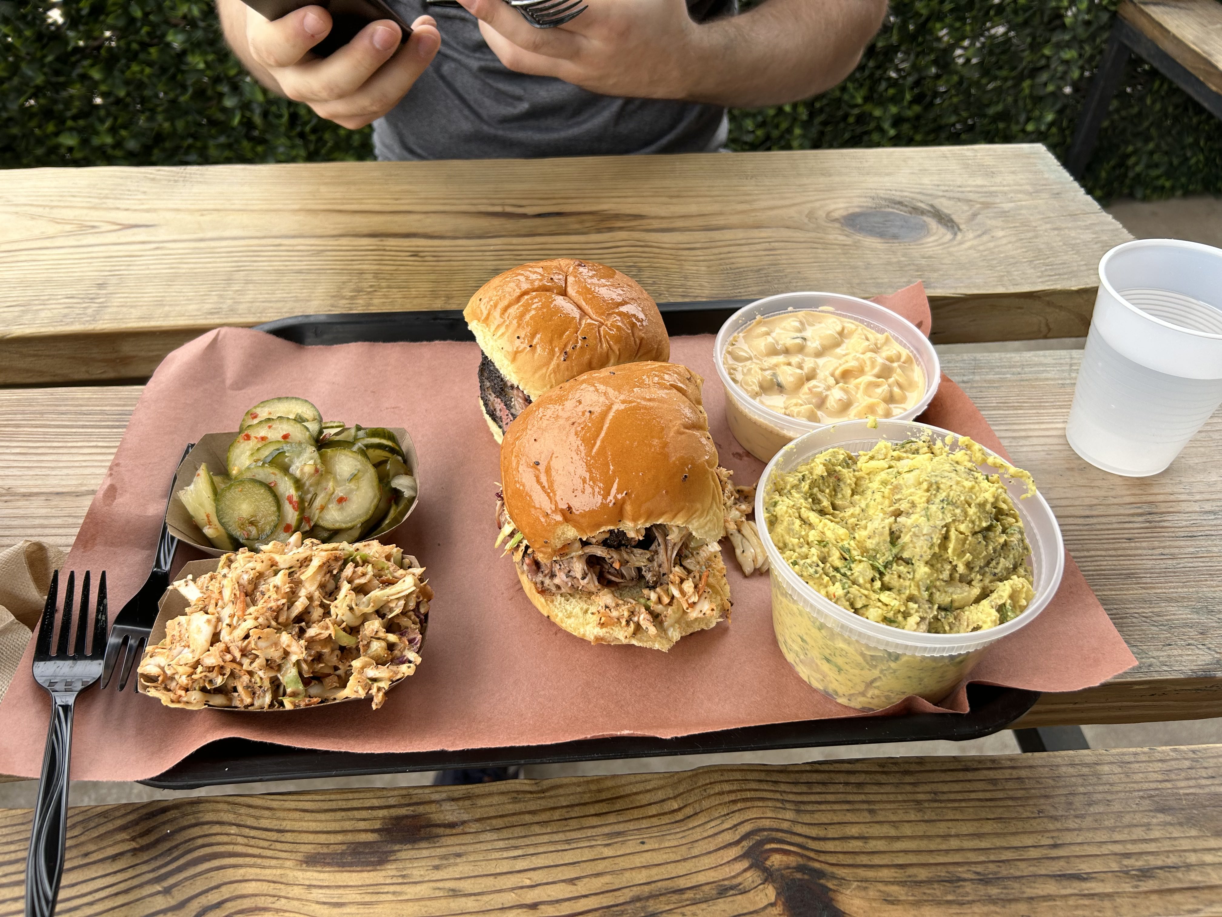 An outdoor feast featuring pulled pork sandwiches, a bowl of macaroni and cheese, potato salad, and pickled cucumbers on a rustic wooden table.