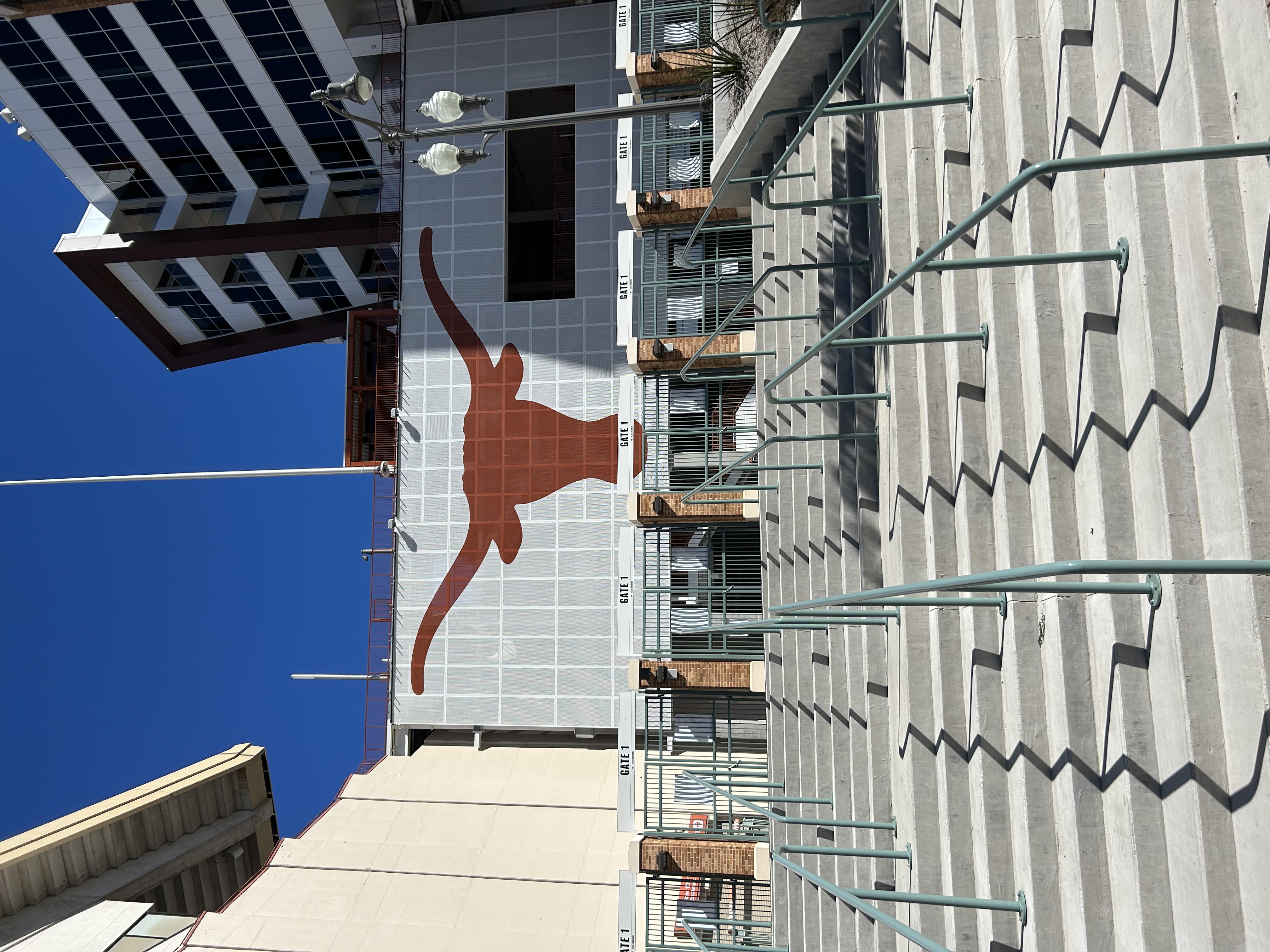 Sunlit steps leading to the entrance of a stadium with a large Longhorn logo, symbolizing the University of Texas, under a clear blue sky.