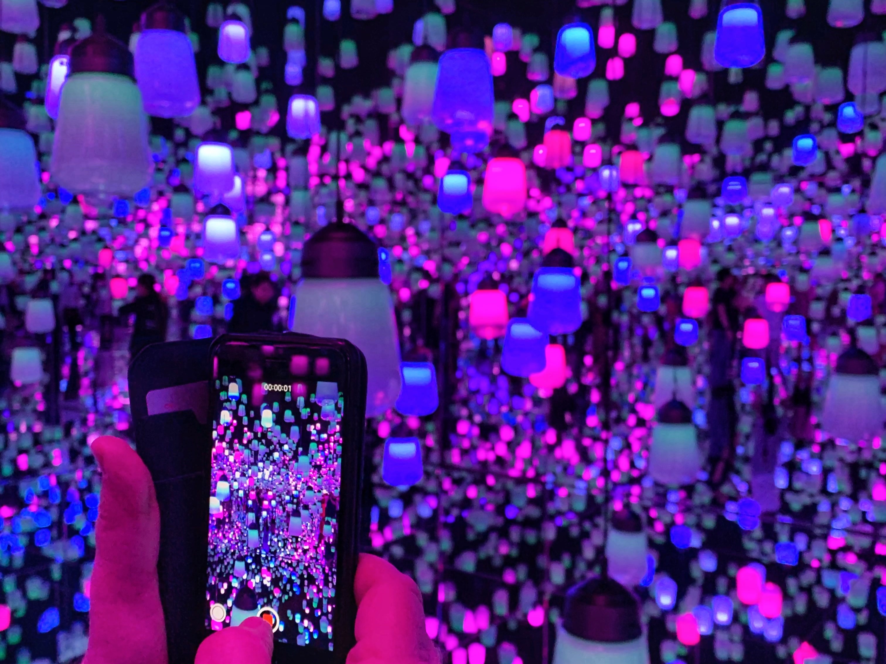 Hands holding a smartphone to photograph multicolored bell lights creating an infinite reflection effect in a mirrored room at teamLAB Planets.