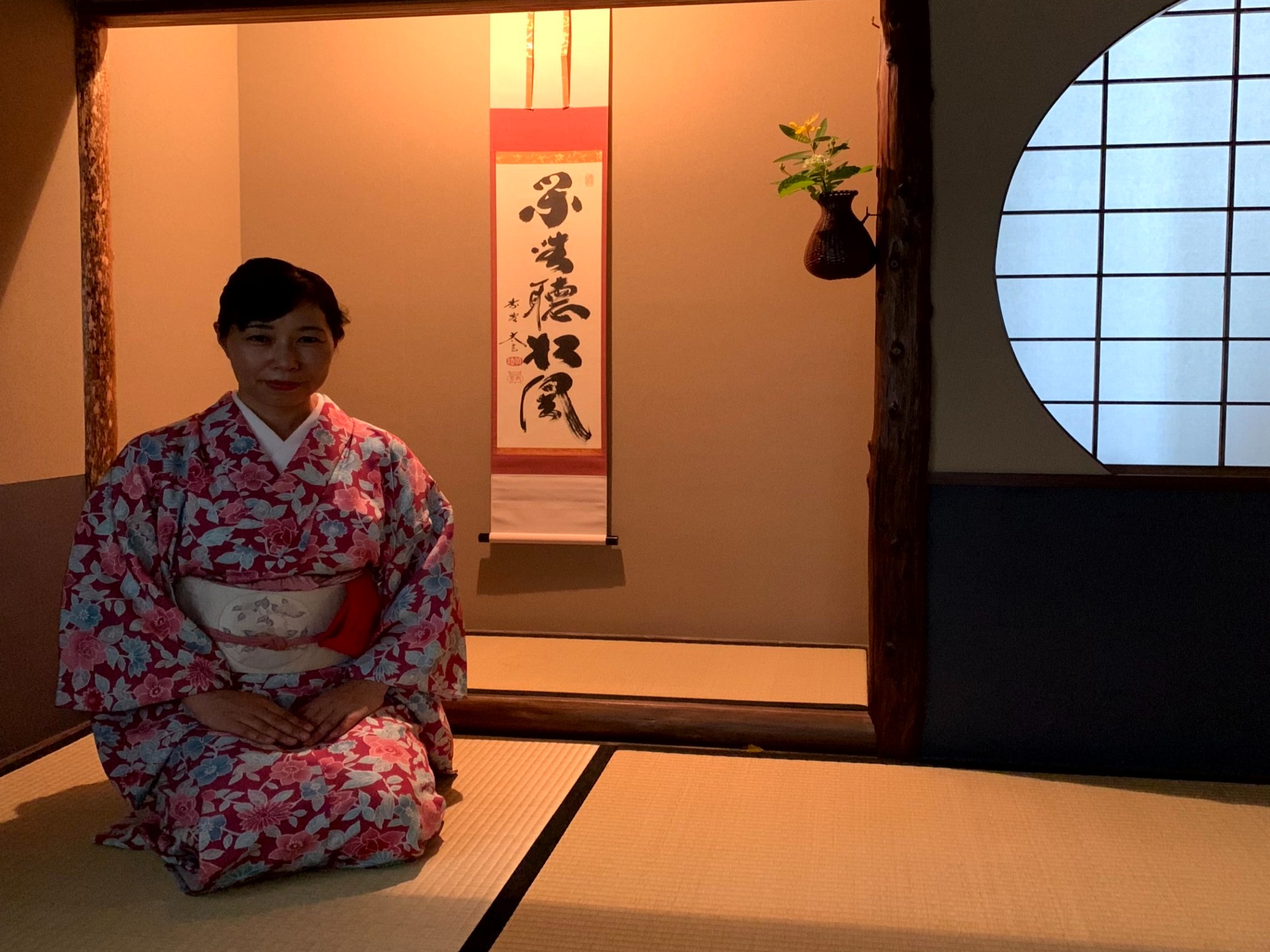 A woman in a floral kimono kneeling in a traditional Japanese room with a red and black wall hanging behind her.