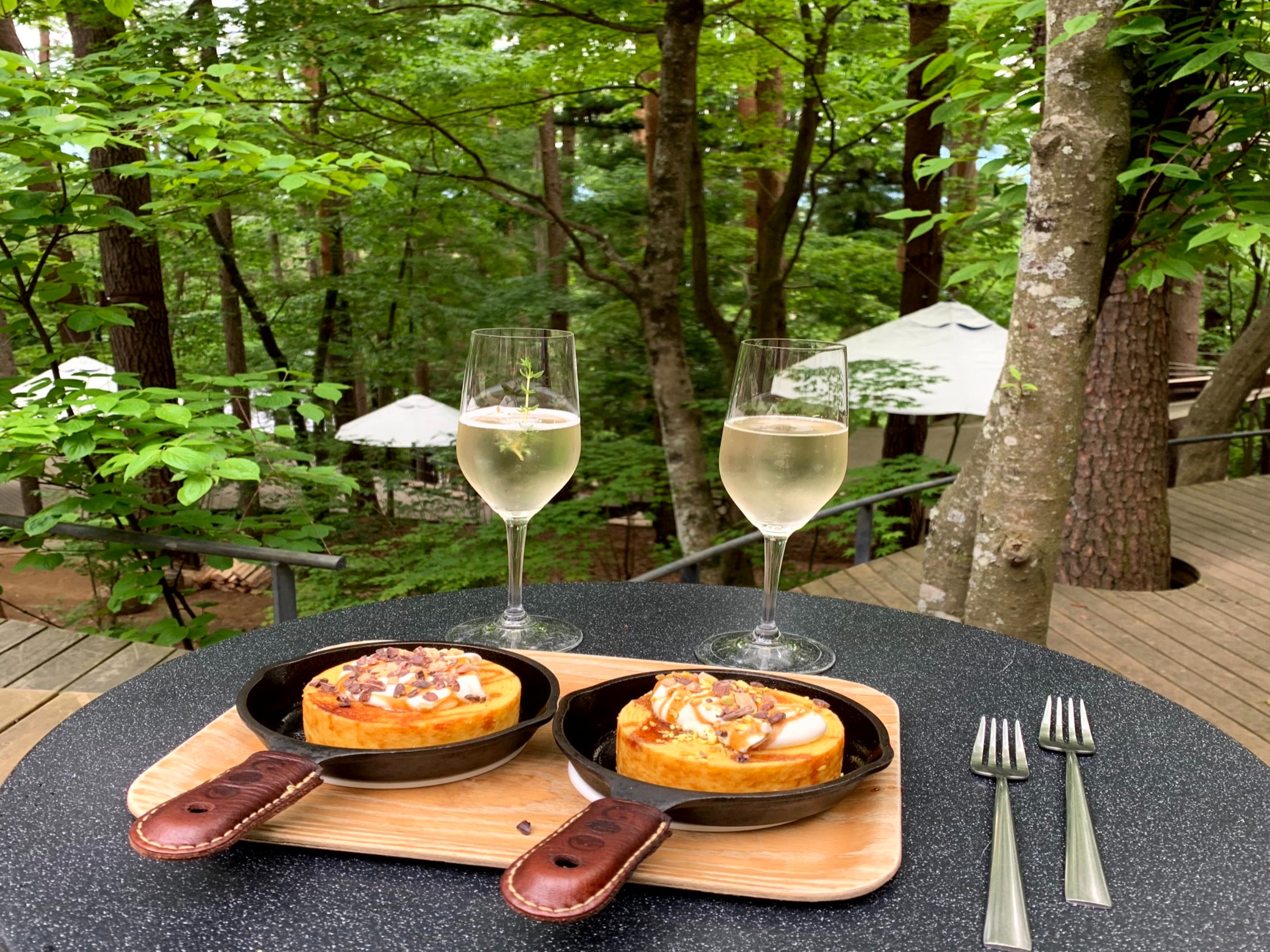 An elegant outdoor dining arrangement with two personal-sized cast iron pans on a wooden board complemented by white wine and set against a backdrop of lush greenery.