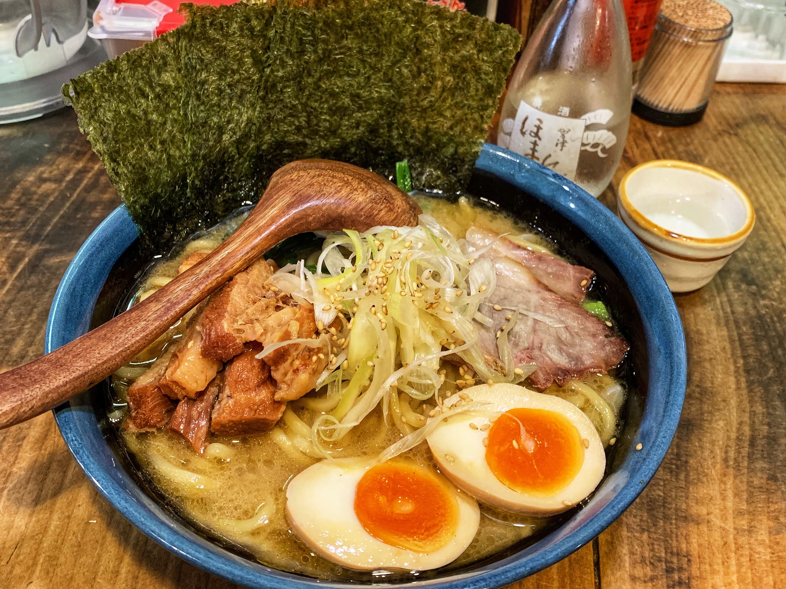 A blue bowl of ramen topped with eggs and meat and a wooden spoon on a wooden table.