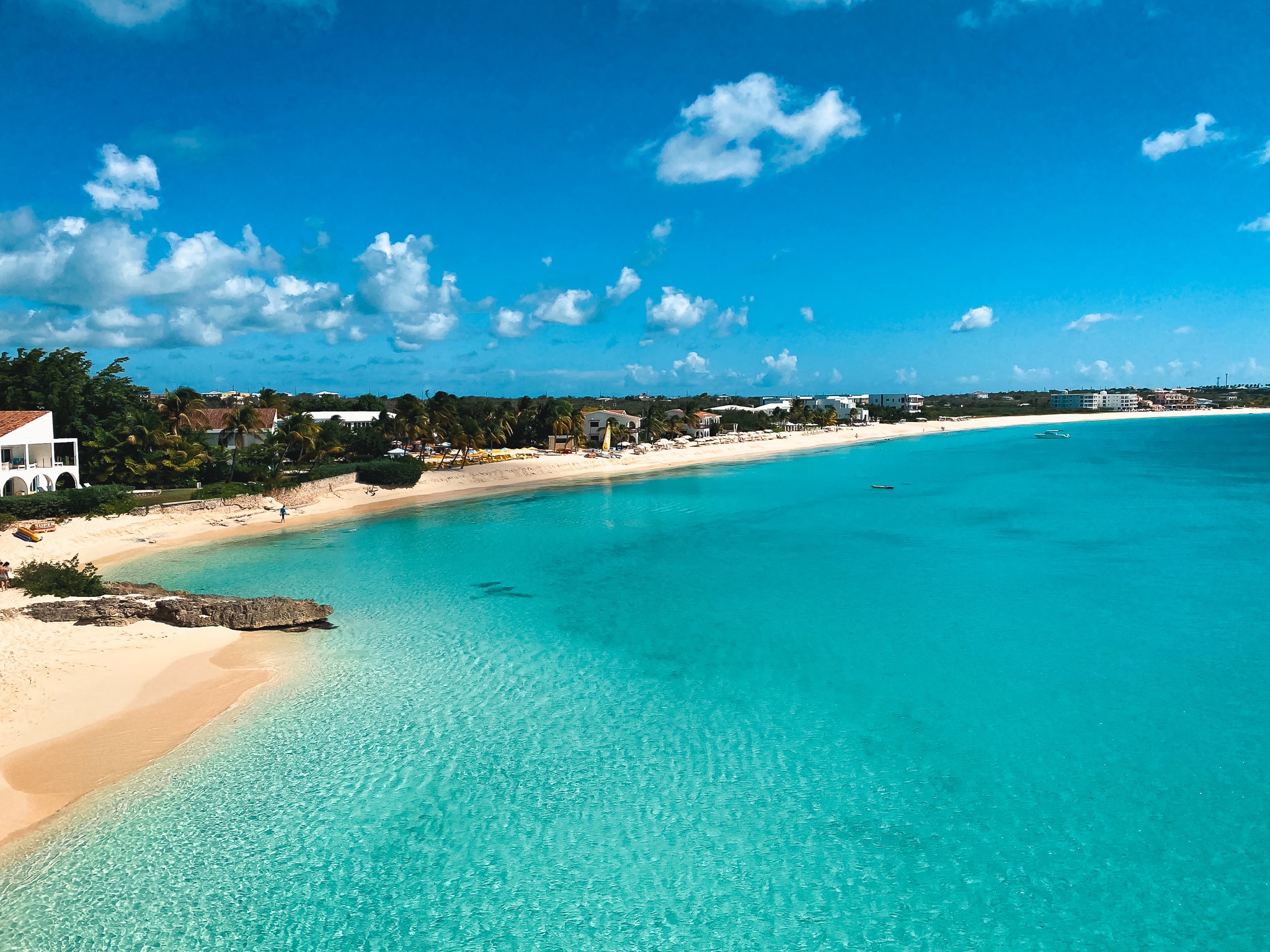 A serene coastal scene where turquoise waters meet a sandy beach, with buildings and greenery under a cloud-dotted blue sky.