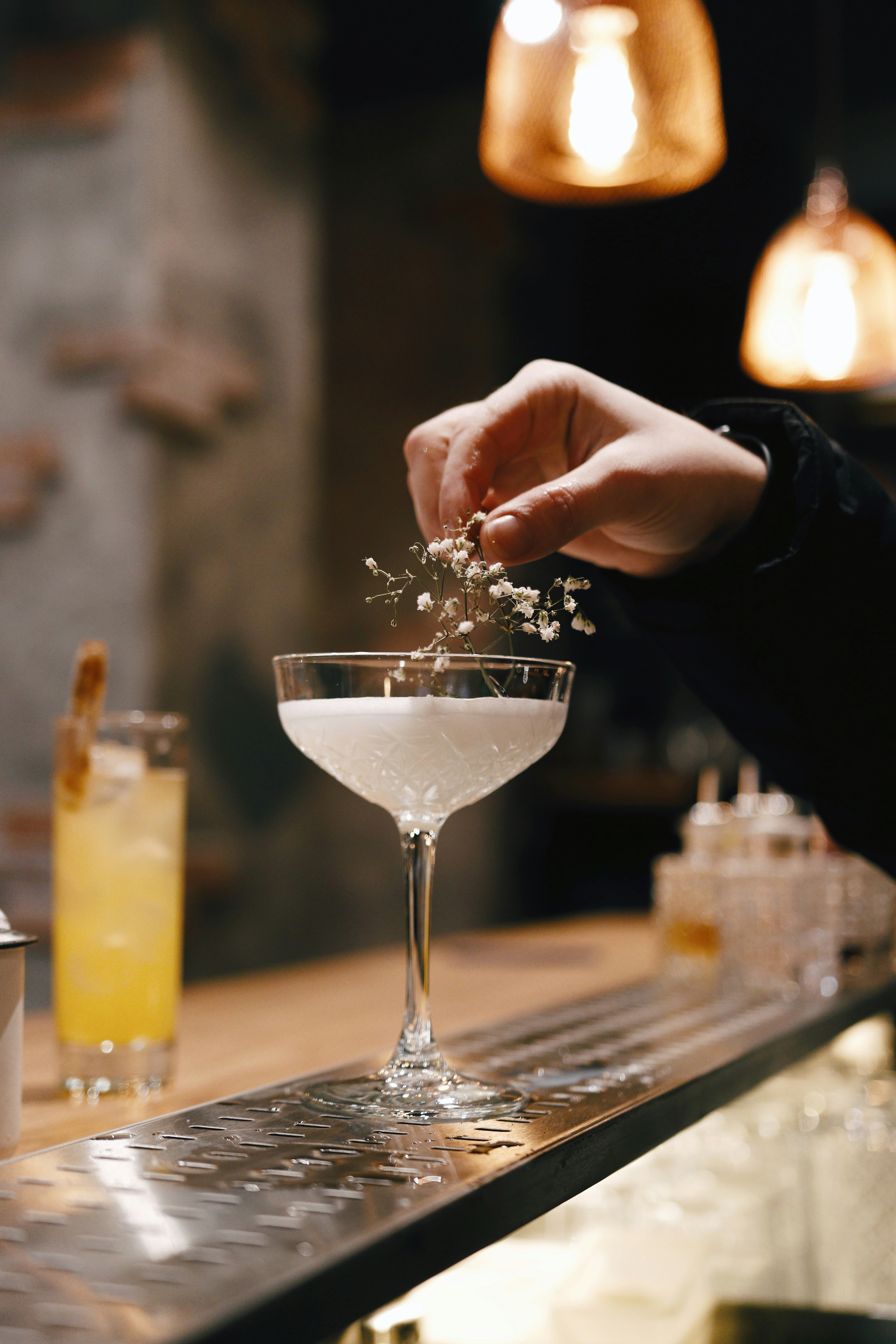 A white cocktail on a bar with a hand adding a sprig to it