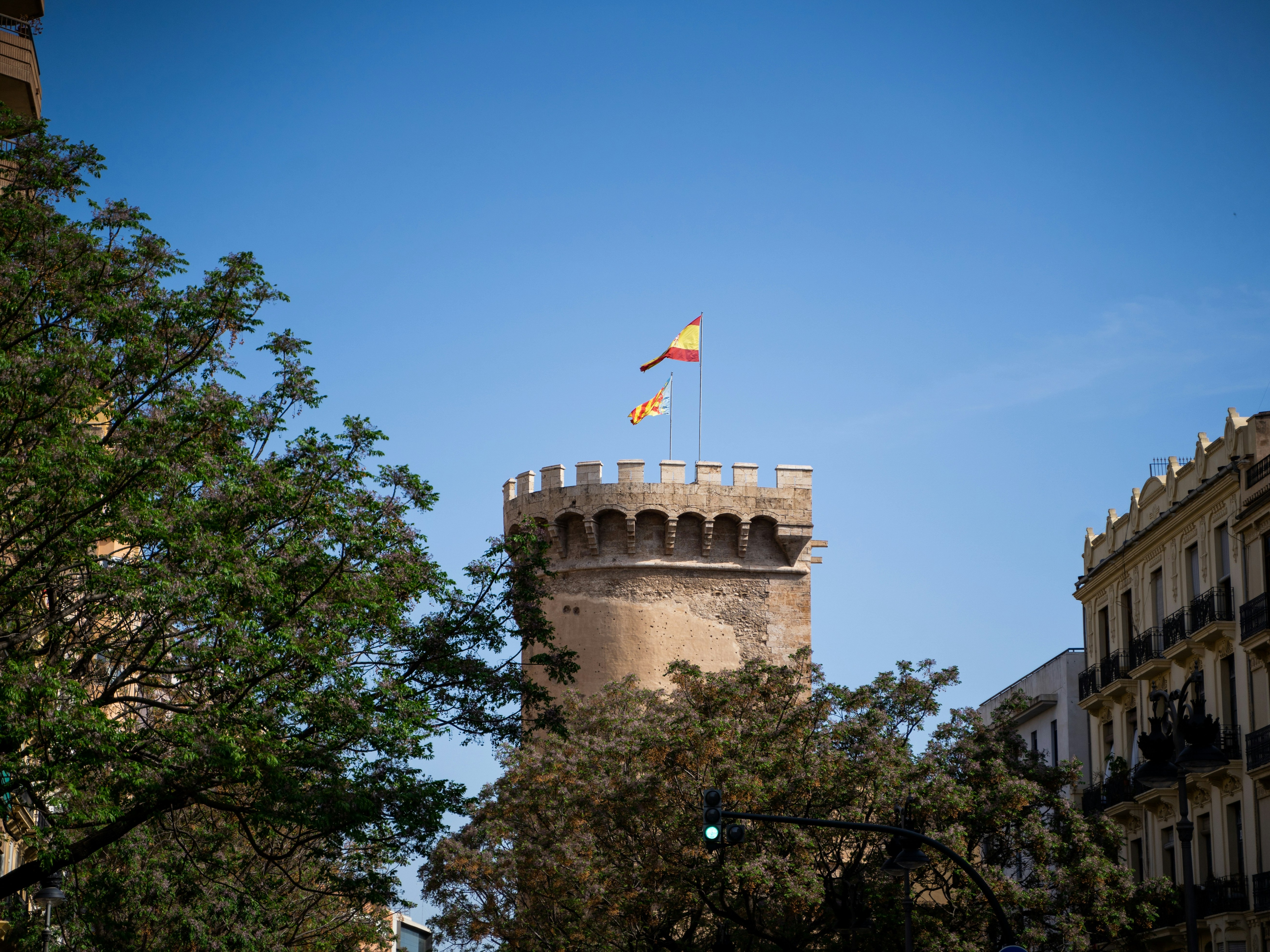 A historic Spanish castle tower with a waving flag, flanked by modern buildings and greenery under a sunny sky.