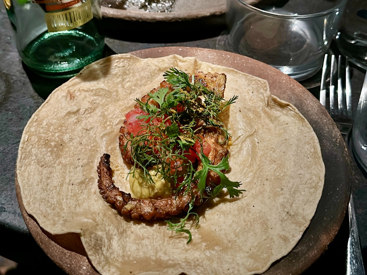 A plate covered in a tortilla, and topped with a piece of grilled octopus, salad, herbs and sauces.