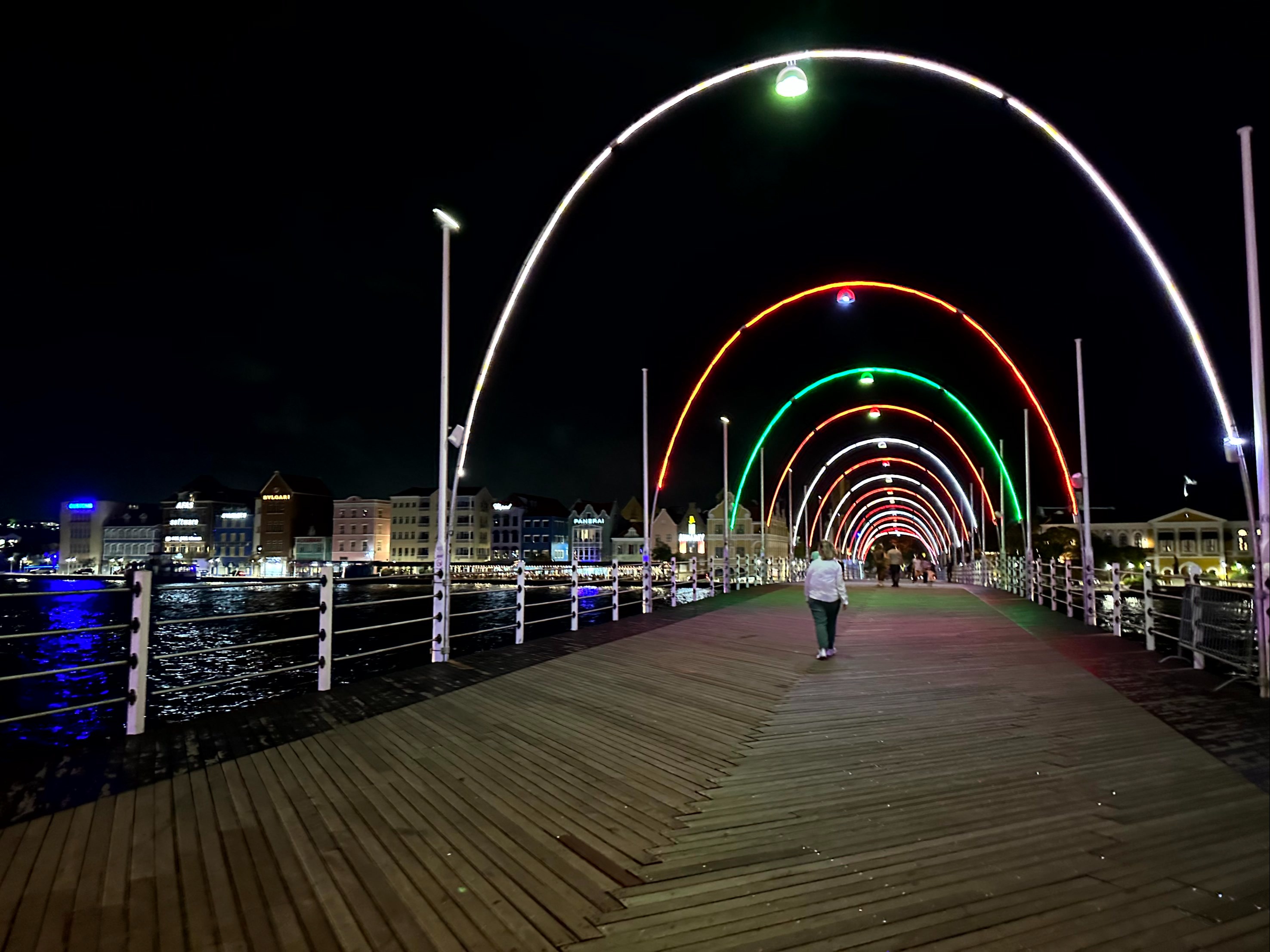 The image captures a solitary figure walking on a vibrantly lit pier at night.