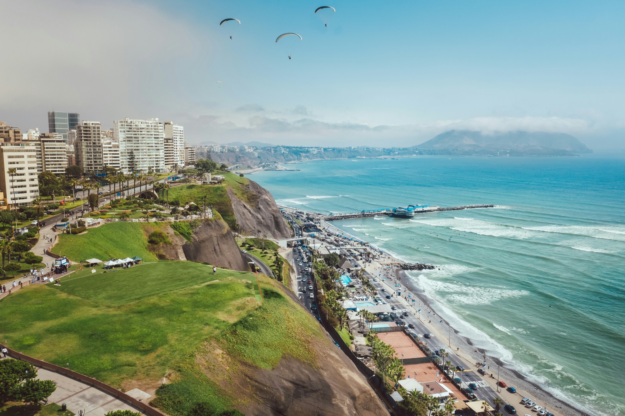 An aerial view of Lima, Peru with the city skyline, coastline, azure water, people parasailing and mountains in the background.