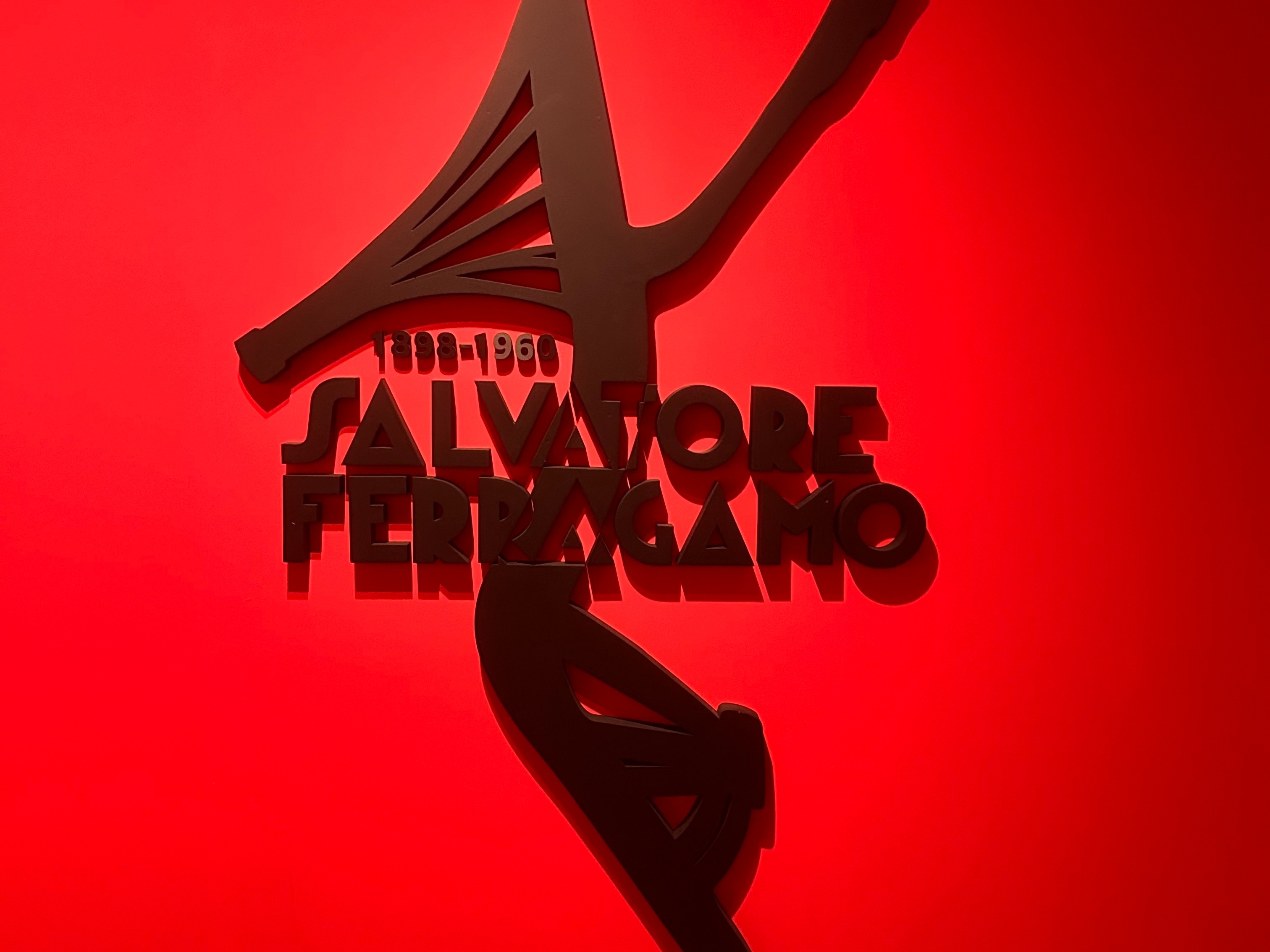 The image showcases a striking Salvatore Ferragamo logo in a metallic finish against a vibrant red backdrop, accompanied by an abstract shoe sculpture.