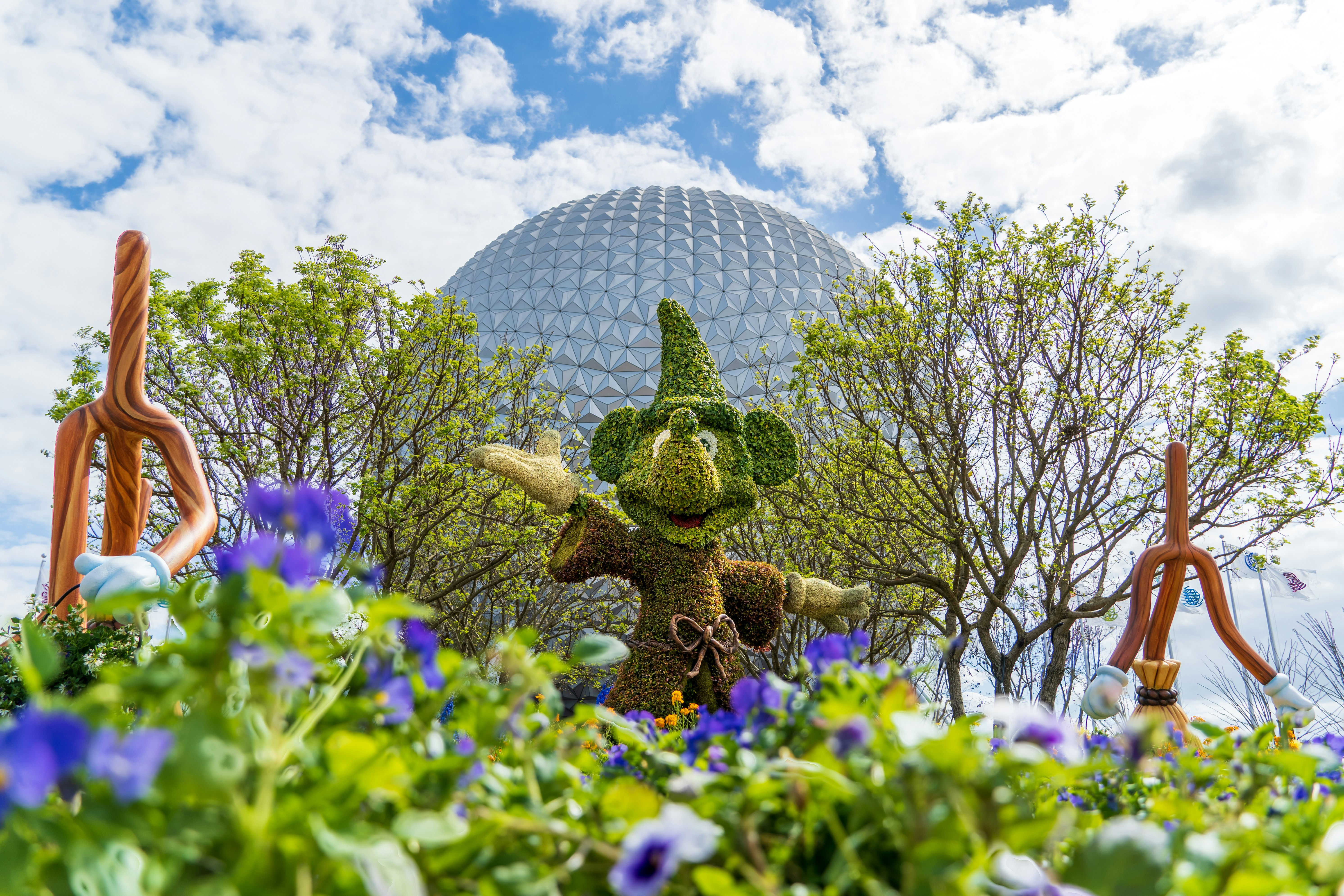 A picture of the famous Epcot sphere with foliage and greenery in front with a Mickey Mouse-shaped bush.