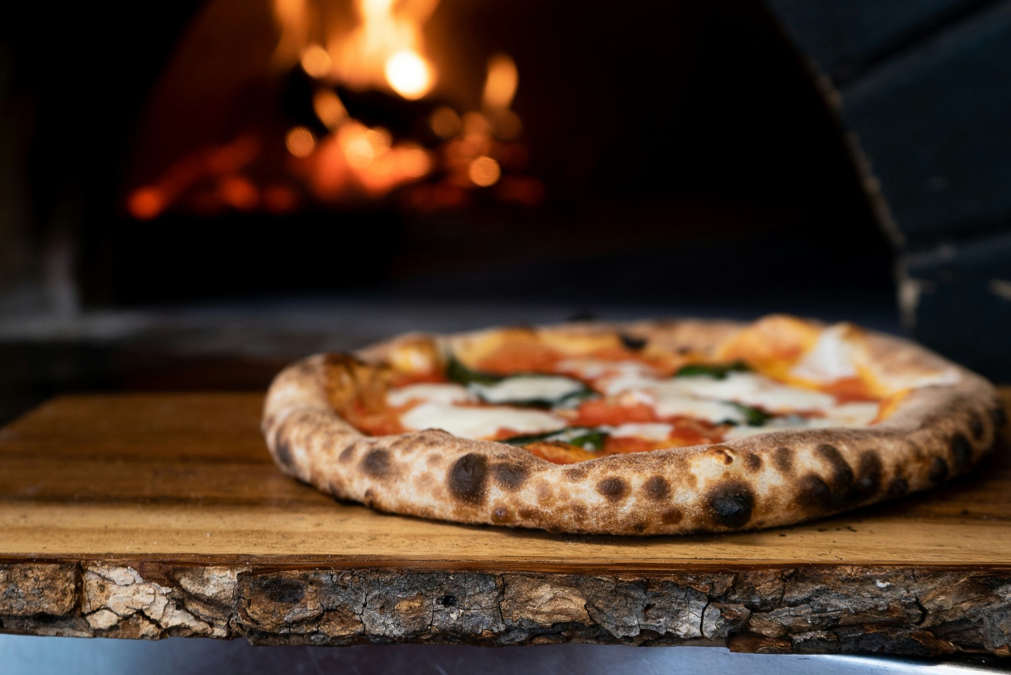 A whole, freshly baked margarita pizza on a wooden board, with a blurred fire of a wood-burning oven in the background.