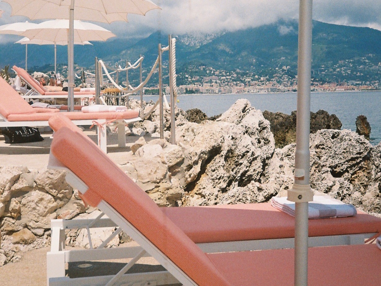 Salmon-colored sun beds, white umbrellas, on a French beach with rocks, sea and mountains in sight.