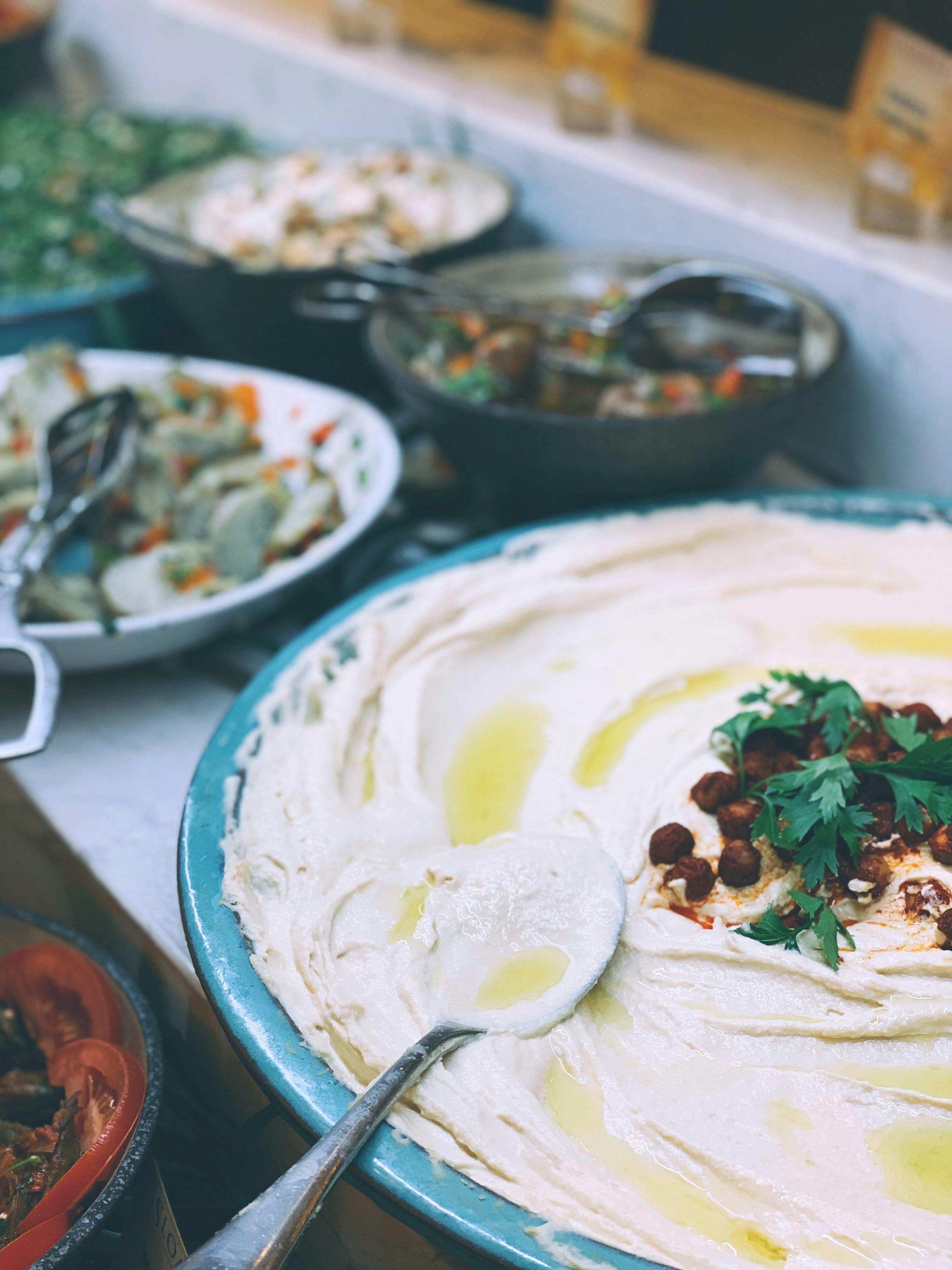 A plate of freshly made hummus, topped with olive oil, herbs and toasted chickpeas, with other salads and dishes blurred in the background.
