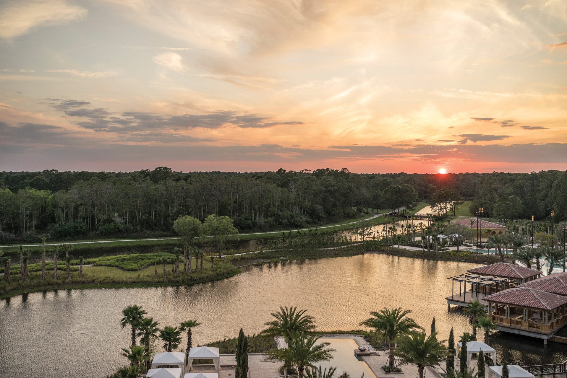 A lake surrounded by overwater bungalow-type buildings, palms, and the resort's well-manicured grounds as the sun sets in the distance