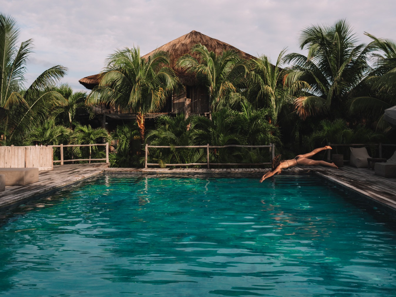 This image depicts a straw roof cabana behind a swimming pool, while also being surrounded by palm trees. There is a person diving head first into the swimming pool from the right side of the image.