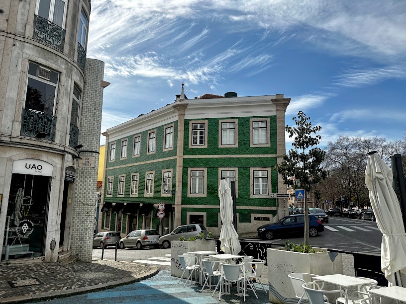 A corner junction of several streets, showing an outdoor dining area of a cafe and a pretty green and white building in the background