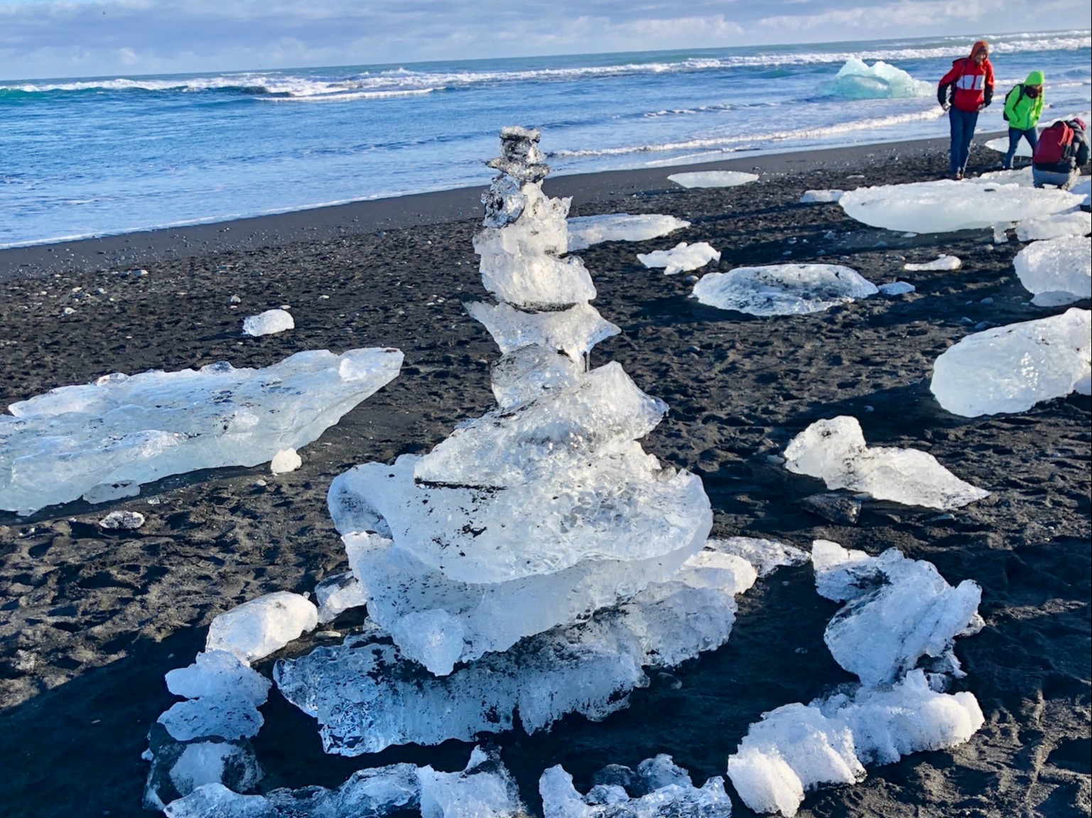 The image depicts a tower of ice chunks on a black sand beach, with the ocean and two people in the background.