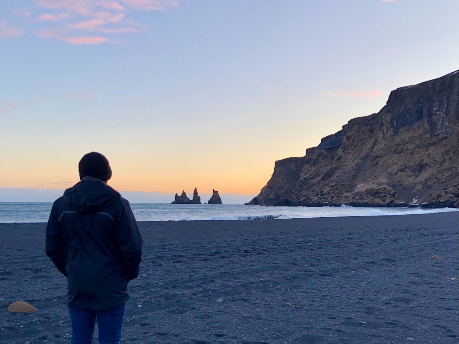 A person standing on the beach looking toward the water at sunset with a rocky hill in the distance