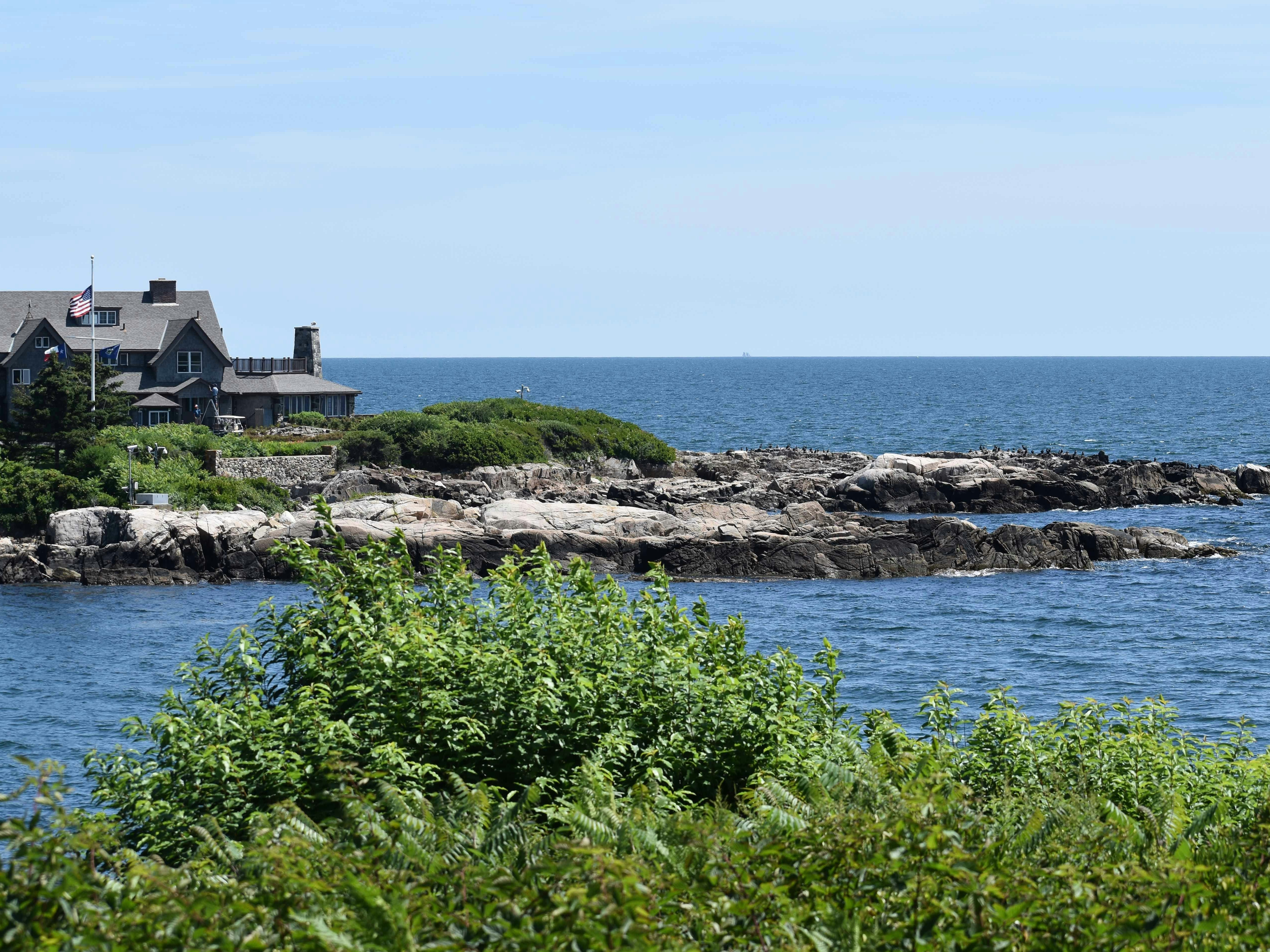 The image captures a serene coastal setting with a house perched on the rocky shore, under a clear blue sky.