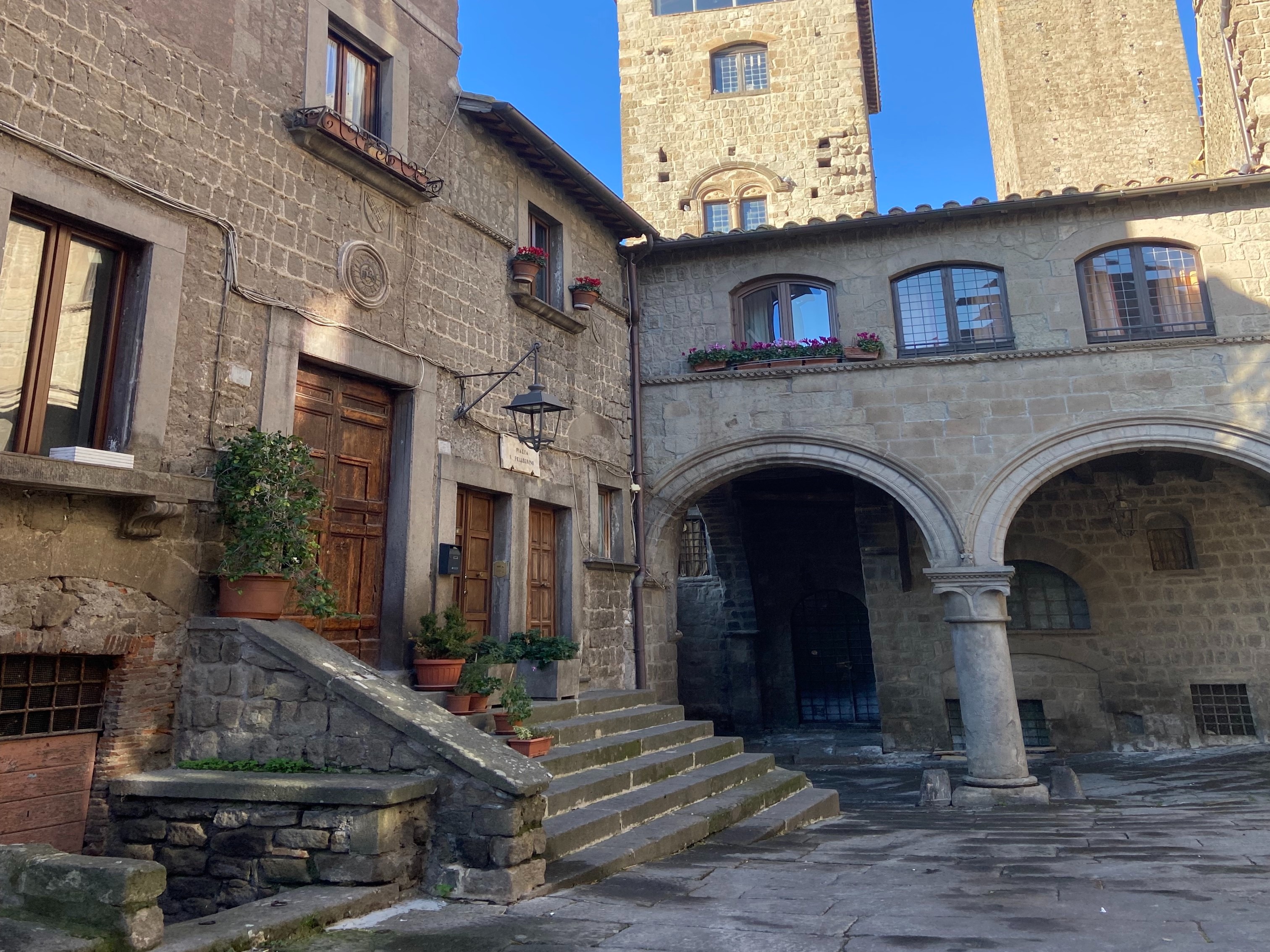 A sunny day in an old European town, showcasing a stone building with wooden shutters and a staircase leading to an arched entryway.