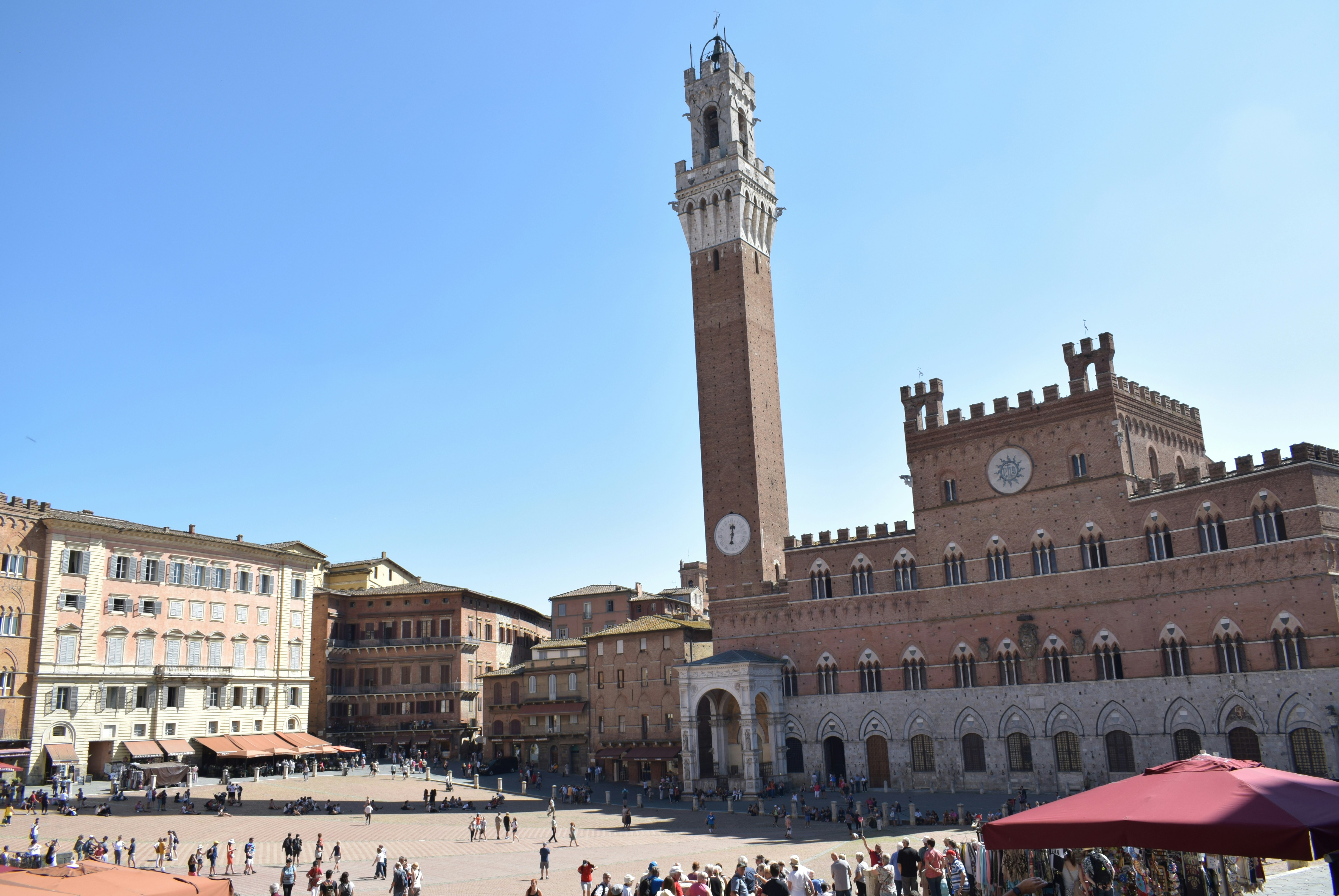 The image captures a bustling public square with a historic clock tower and surrounding buildings under a clear blue sky.