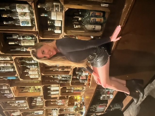 Chelsea in a black top and leather mini skirt sitting on a bar in front of bottles of alcohol.
