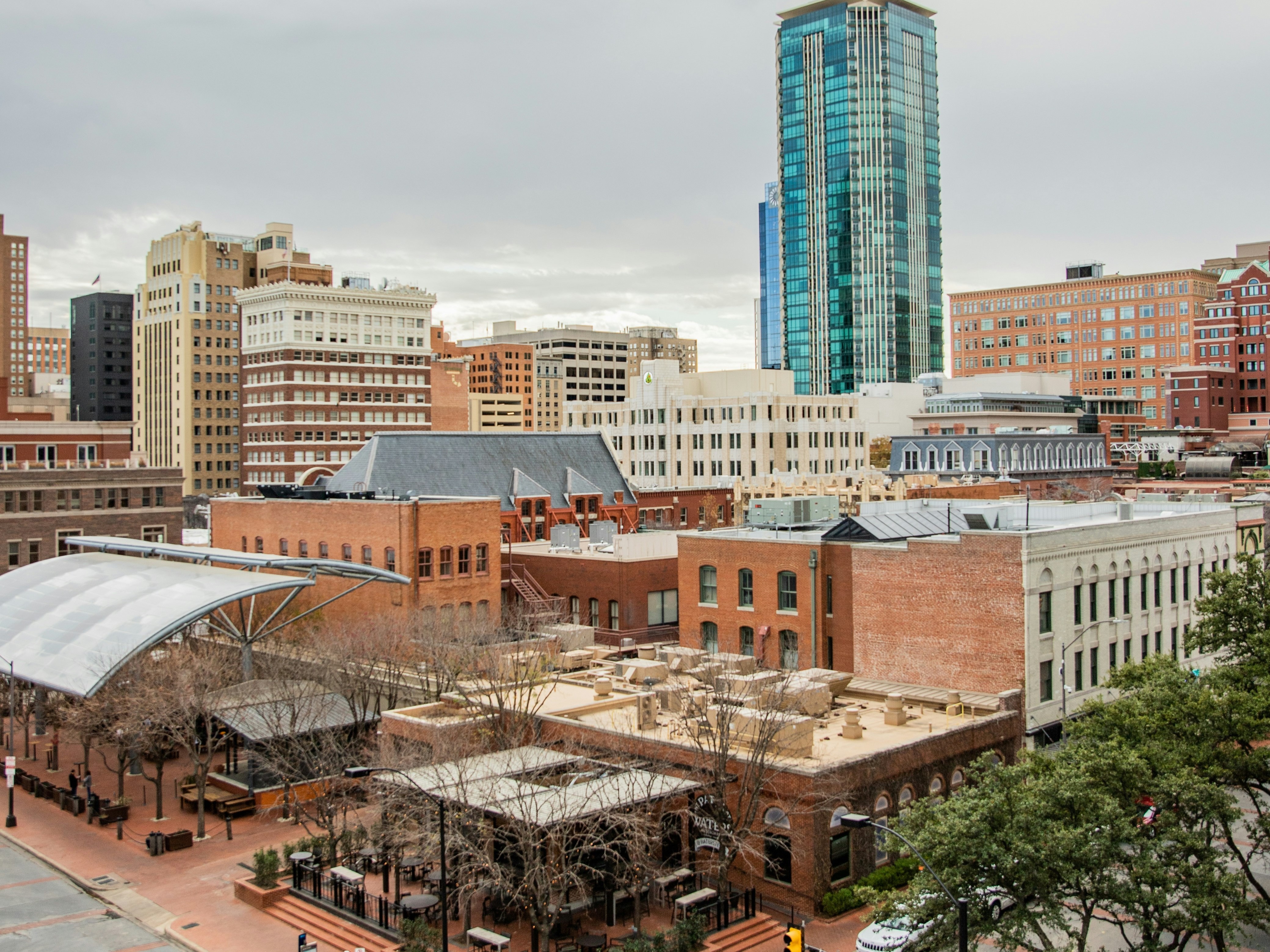 The image depicts a cityscape with a blend of modern and traditional architecture, a plaza, and a tall glass skyscraper under a cloudy sky.