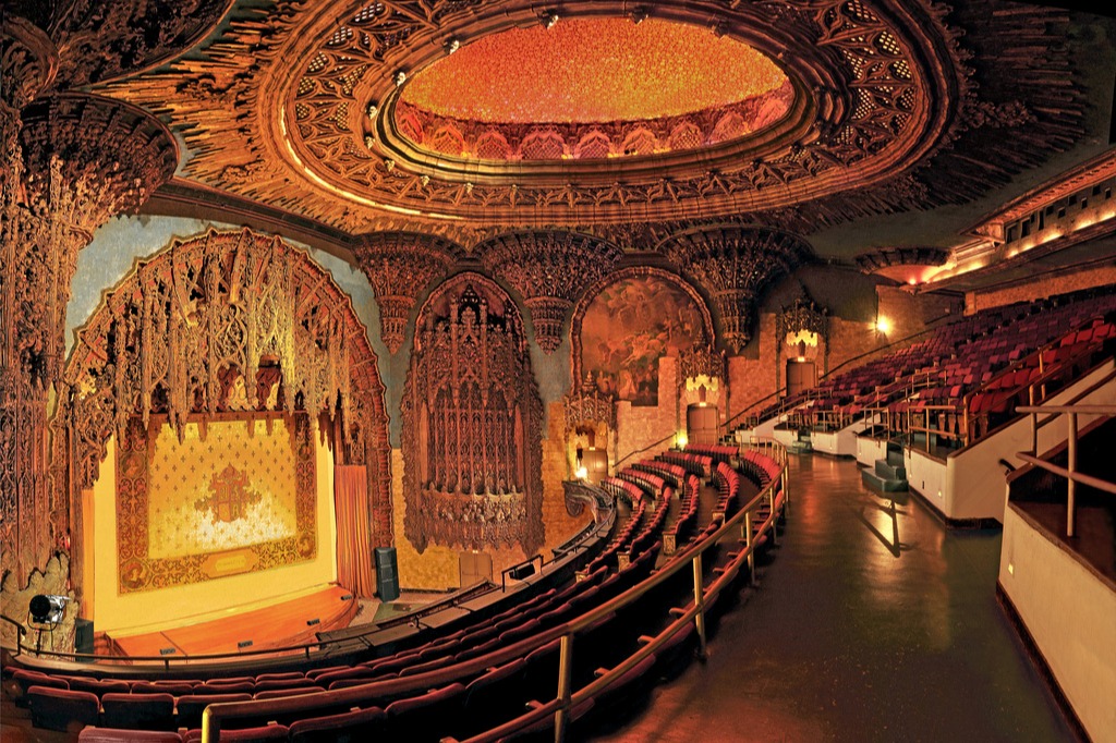 This image depicts an old theatre with incredible stonework, layers of seats, an organ player, decorative ceiling, and golden stage. The entire room is lit with warm toned lighting and grand looking.