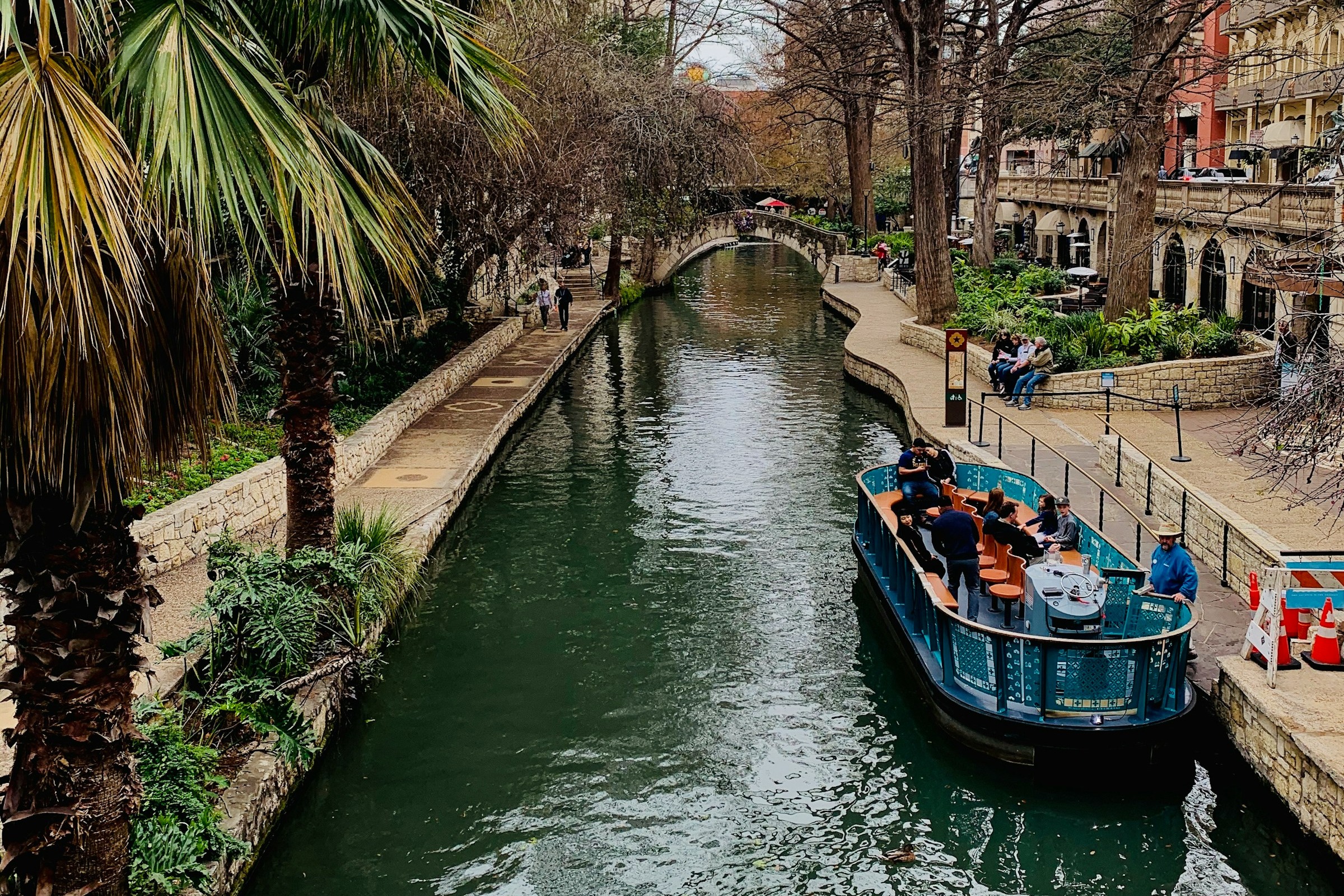 A local boat ride, on an open, blue boat along the Arneson River, bordered by palm trees, shops, architecture and people in the distance.