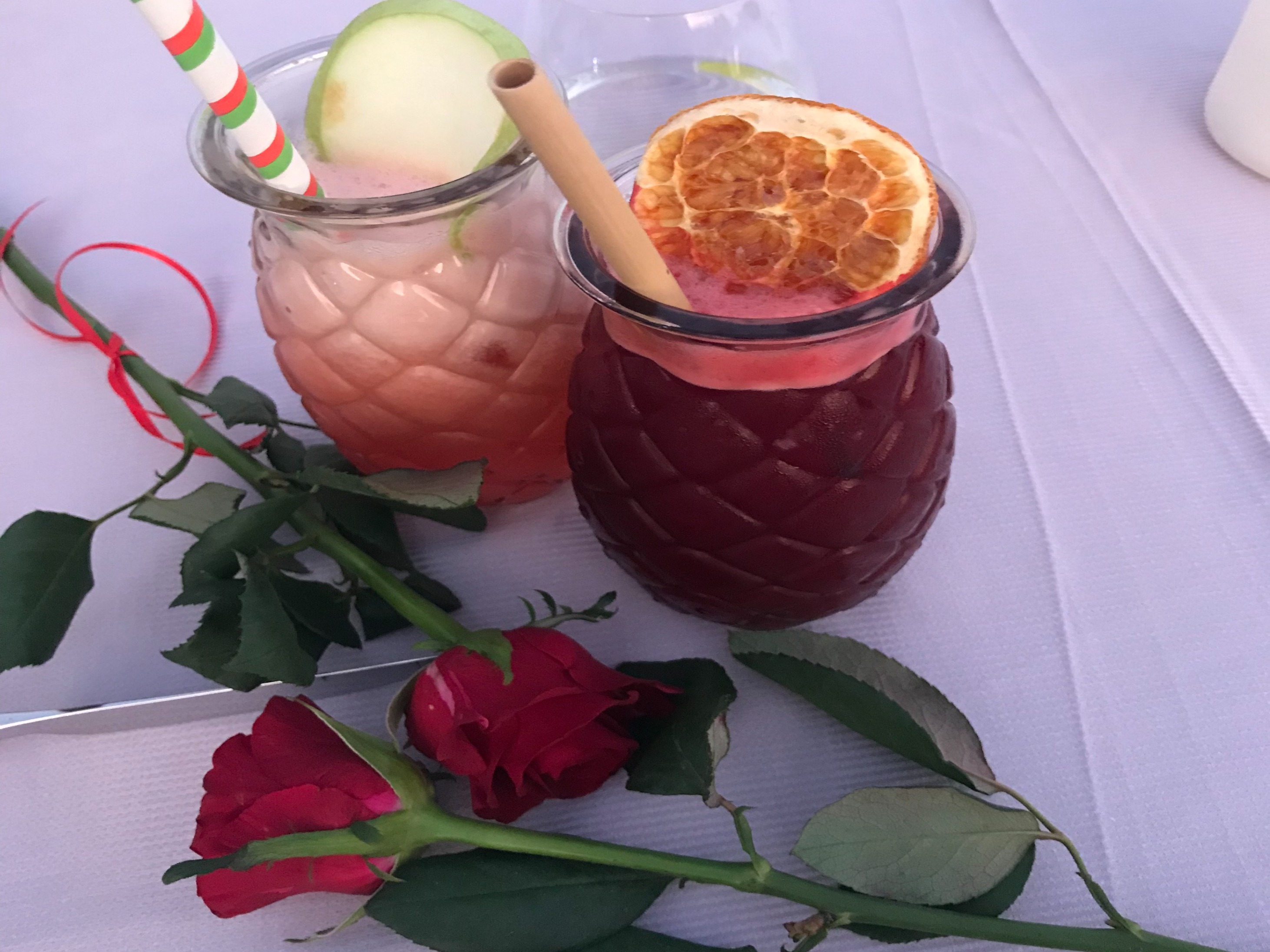 Two cocktails in fancy glasses with fruit toppings next to red roses on a white tablecloth.