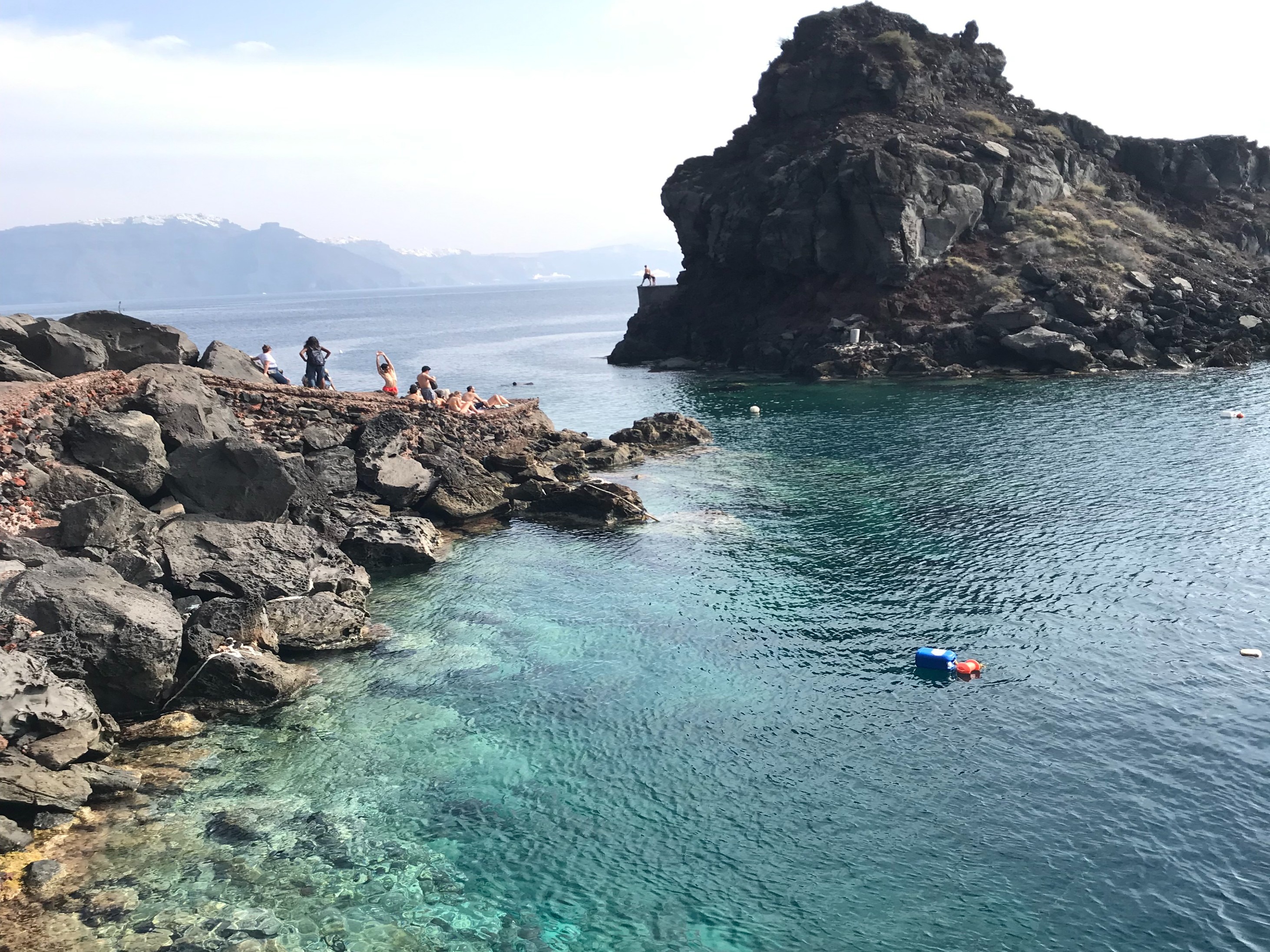 A serene coastal landscape, where the clear turquoise waters meet rugged cliffs, with people in swimsuits sitting on rocks.