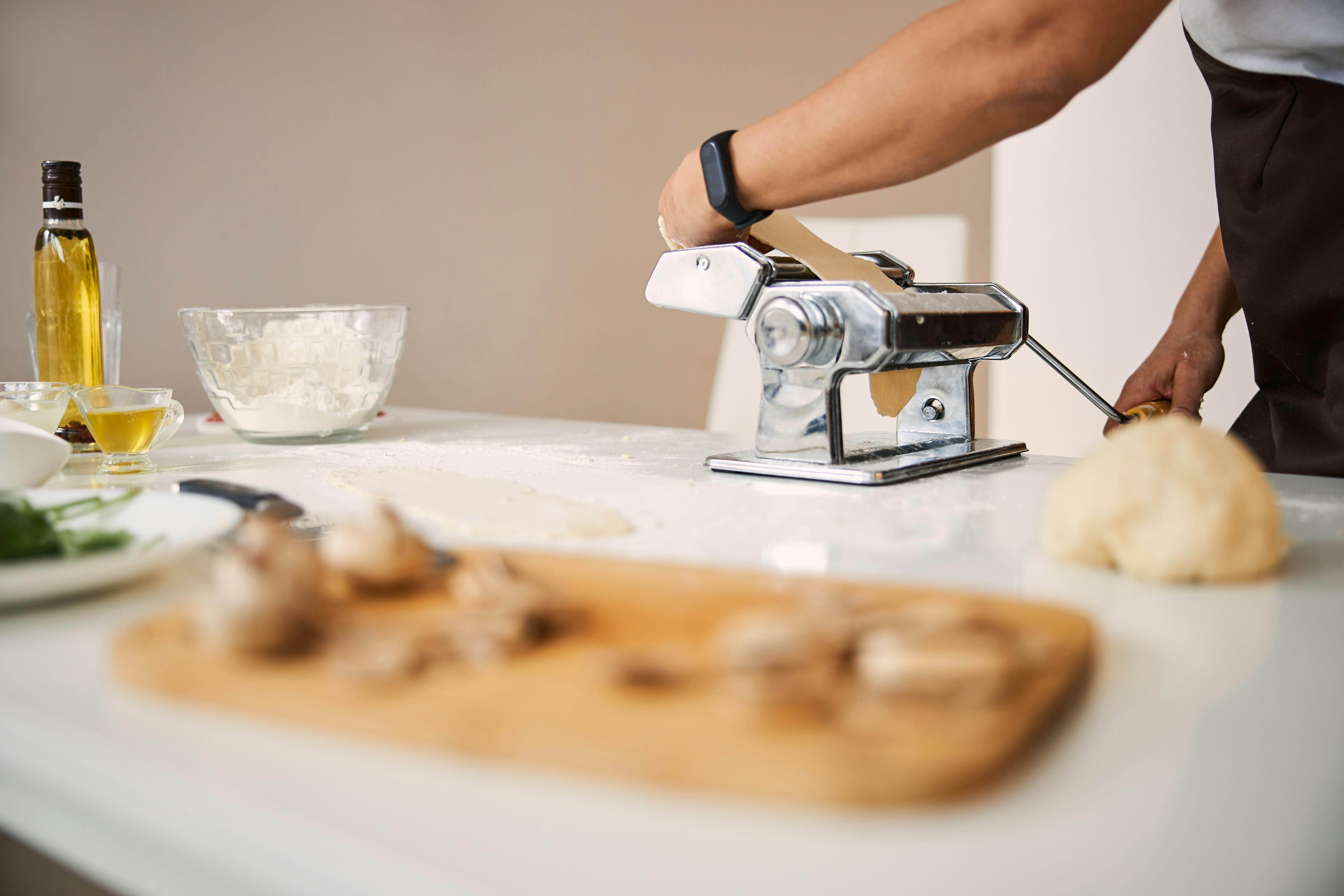 The image showcases a homemade pasta-making session, with hands operating a pasta machine amidst various ingredients on a kitchen counter.