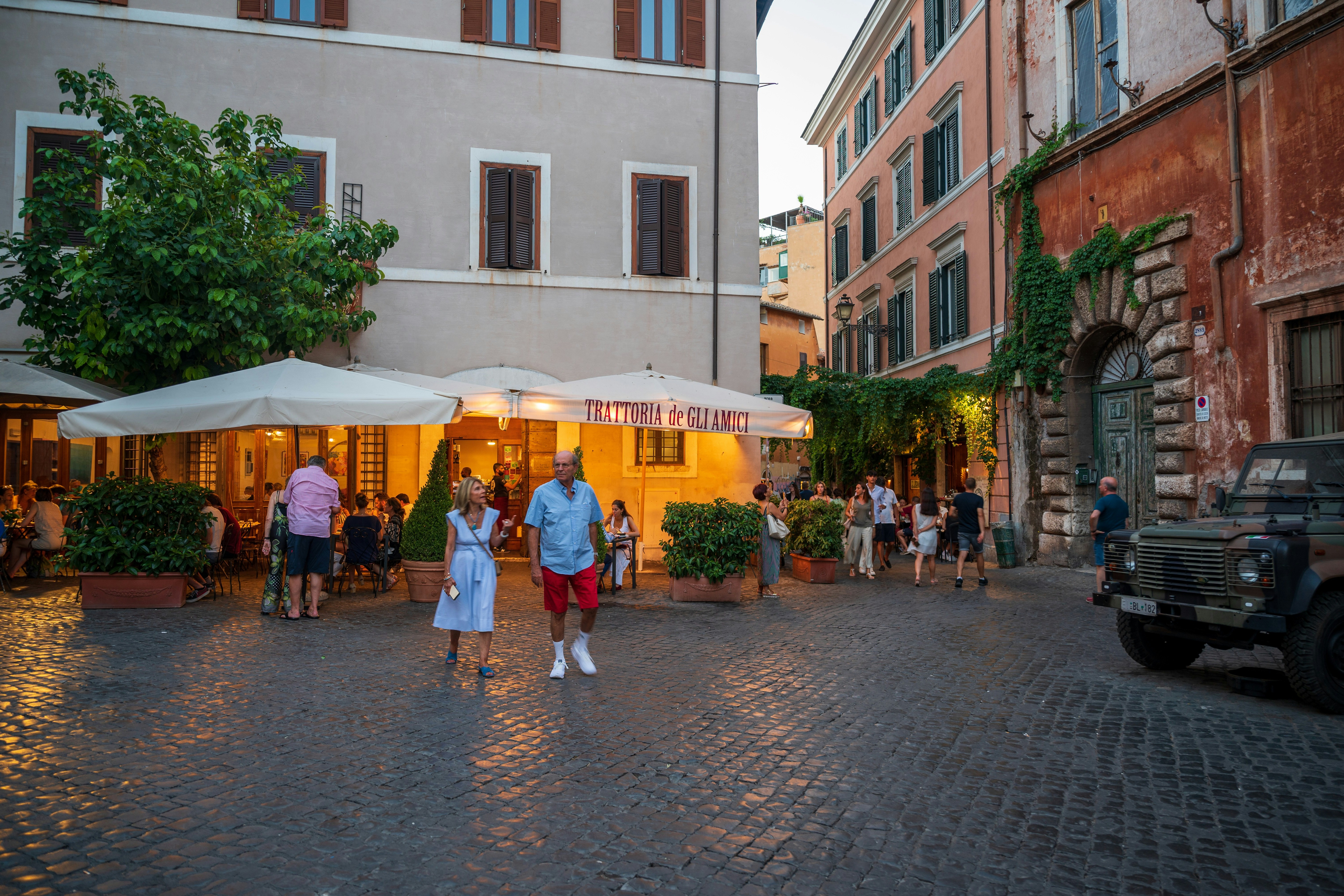 The image captures a vibrant street scene at dusk outside an Italian restaurant, with people enjoying outdoor dining under white umbrellas.