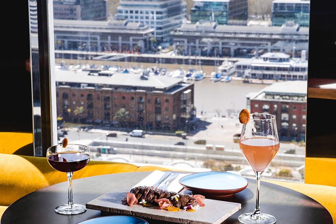 This image depicts a table with two cocktails placed on top of it, yellow seating, a plate with luxurious food on top of it and a view of a city scape in the background.