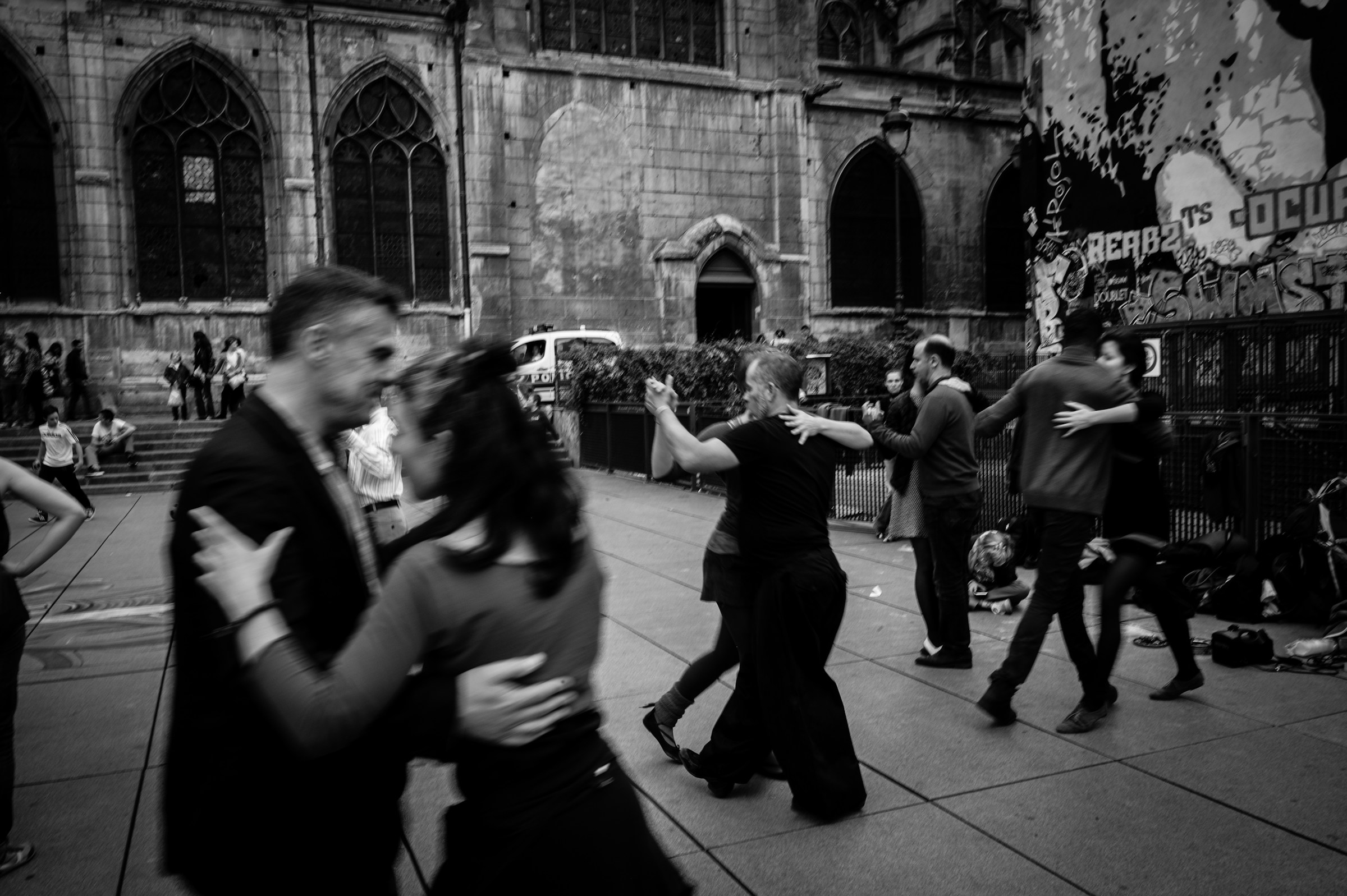A black and white image of people dancing together in an open space with stone walls and arched windows in the background.