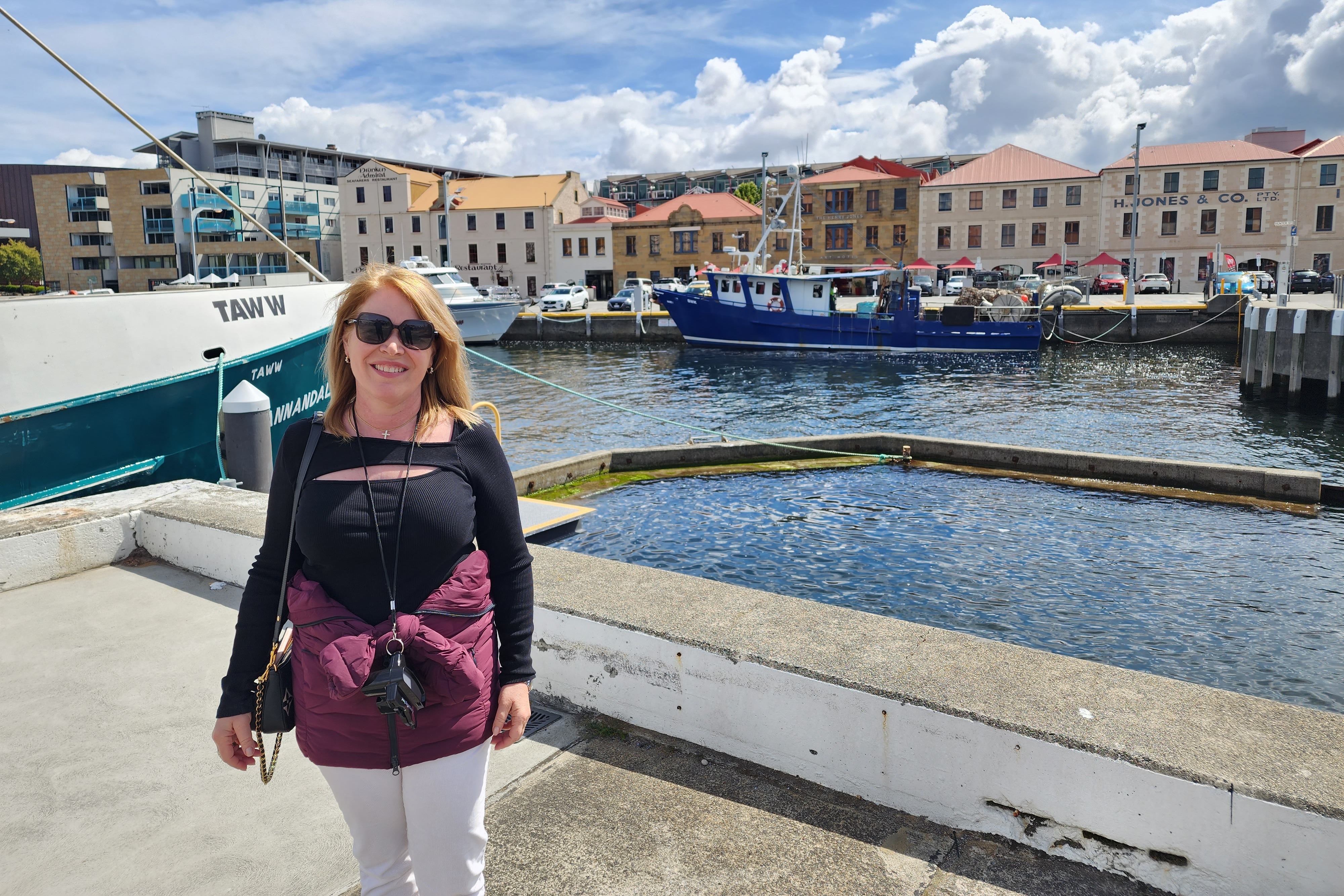 Fora advisor in a black top with a camera around her neck standing in front of a harbor with buildings int he background on a Hobart MacQ1 Tour