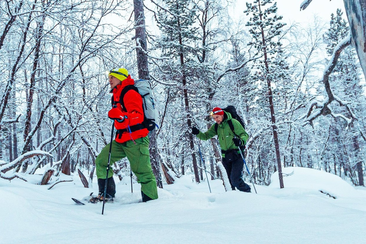 Two people snowshoeing throughout a snowy forest. One person is wearing a yellow hat, red jacket and green pants. The person on the right is wearing a red hat, green jacket and black pants.