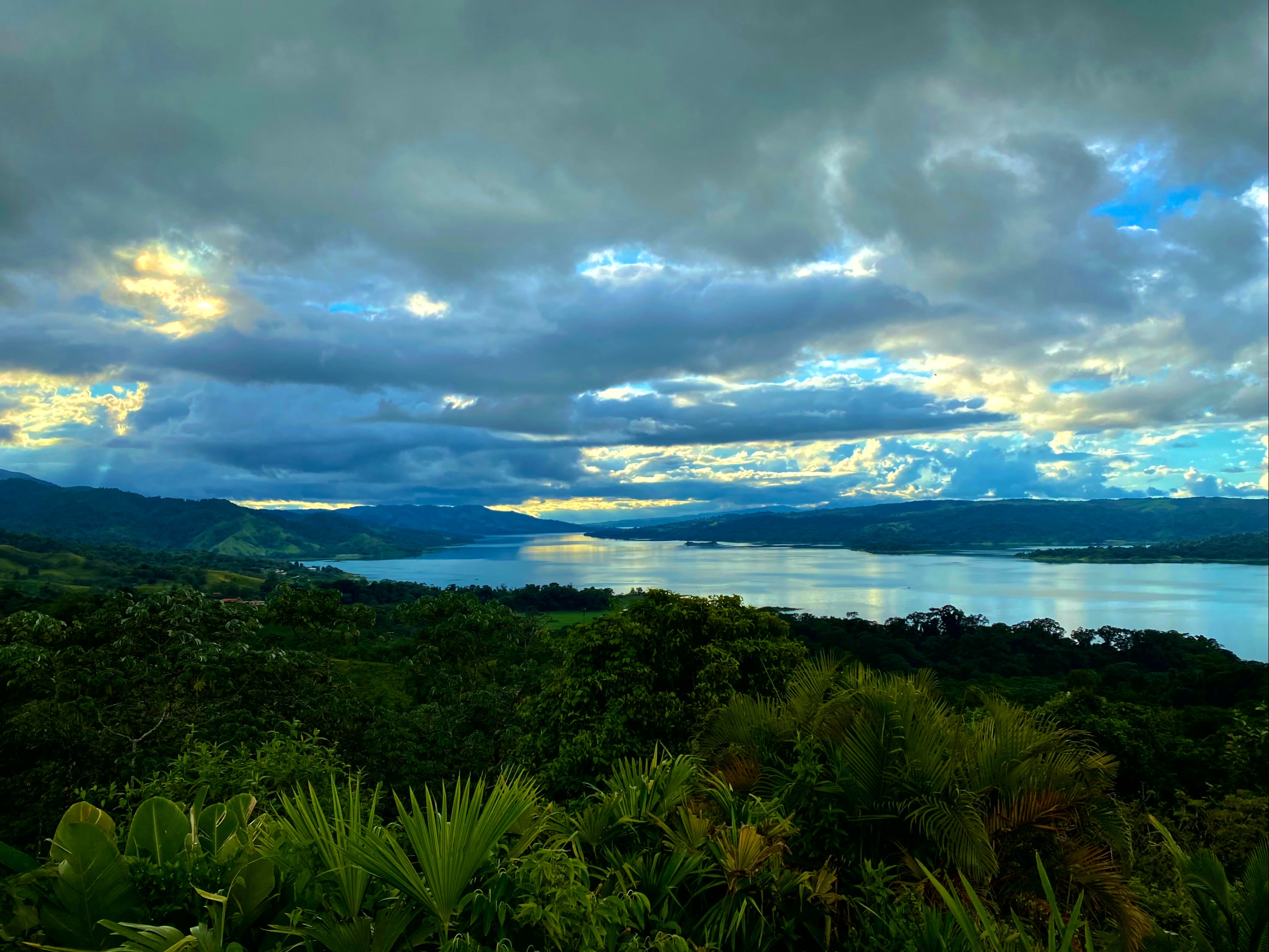 The image depicts a tranquil lakeside landscape with lush foliage and a dramatic, cloud-streaked sky.