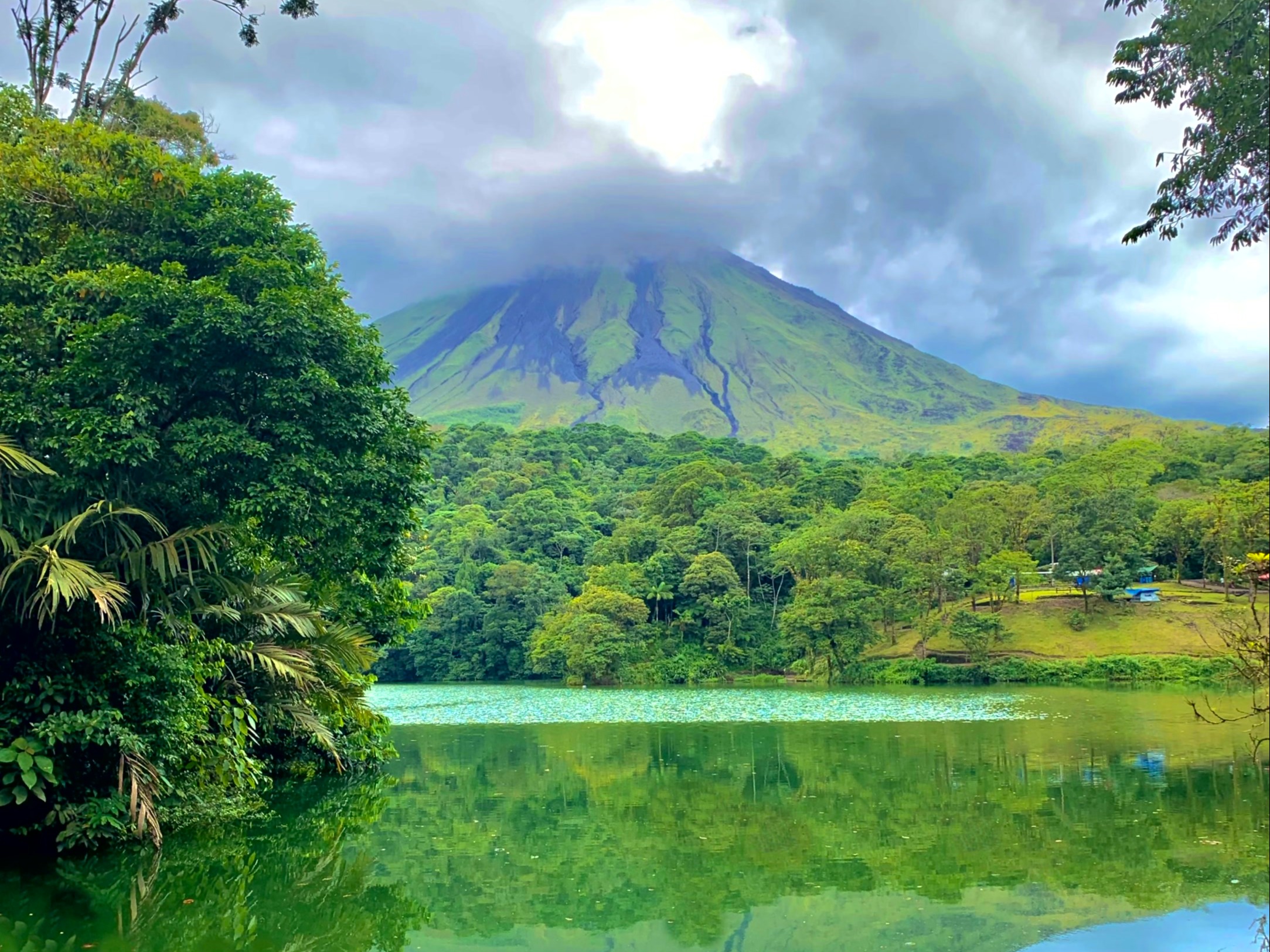 The image portrays a peaceful volcanic mountain with a reflective lake, surrounded by lush greenery under a partly cloudy sky.